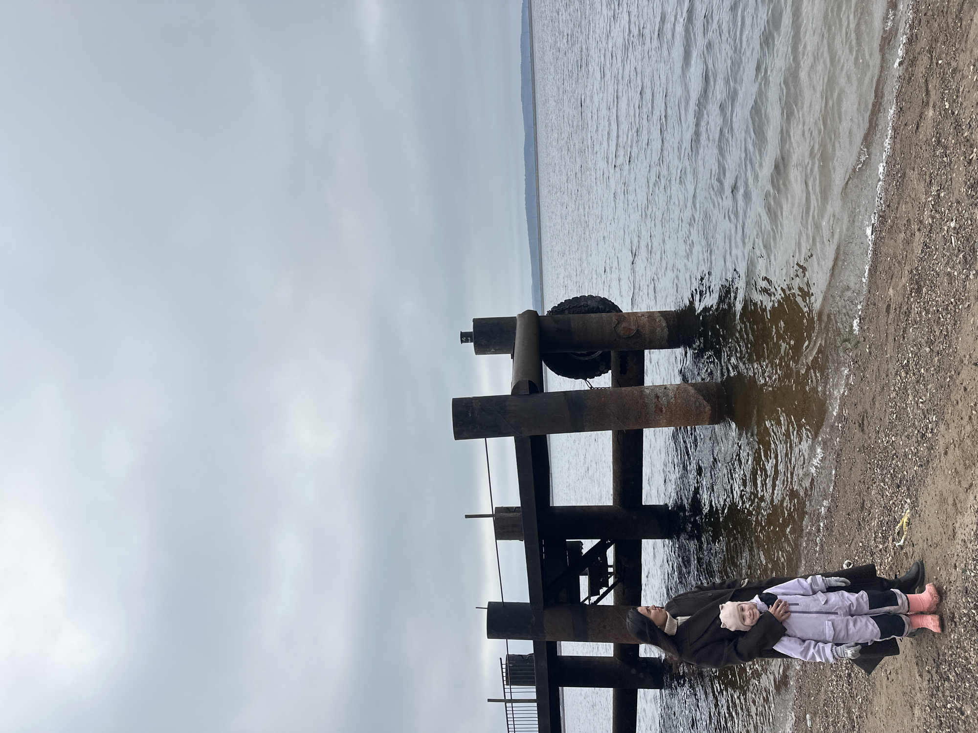 The image captures a serene, overcast day at a waterfront in Chaikovsky, Russia. In the foreground, a woman holds a young child close to her on a pebbly shore. The woman, positioned on the right side of the frame, wears a dark, long coat and a light-colored turtleneck, with her dark hair cascading down. The child, held in her arms, is dressed in a pale purple or grey snowsuit, a light pink hat, and pink boots, looking towards the viewer with a neutral to slightly smiling expression.
To their left and extending into the water is a robust, somewhat dilapidated pier or dock structure made of large, dark-colored beams or piles. One prominent vertical beam in the middle of the structure has a black tire attached to it, possibly for cushioning boats. The structure appears metallic or heavily weathered wood.
The body of water, likely the Kama Reservoir, stretches across the middle and right side of the image, exhibiting choppy, greyish waves. A faint, distant landmass or tree line is visible along the horizon where the water meets the sky. The sky itself is a uniform, light grey, indicating an overcast day without direct sunlight. The overall atmosphere is calm and quiet, possibly cool, given the heavy clothing worn by the individuals. No visible text is present in the image.