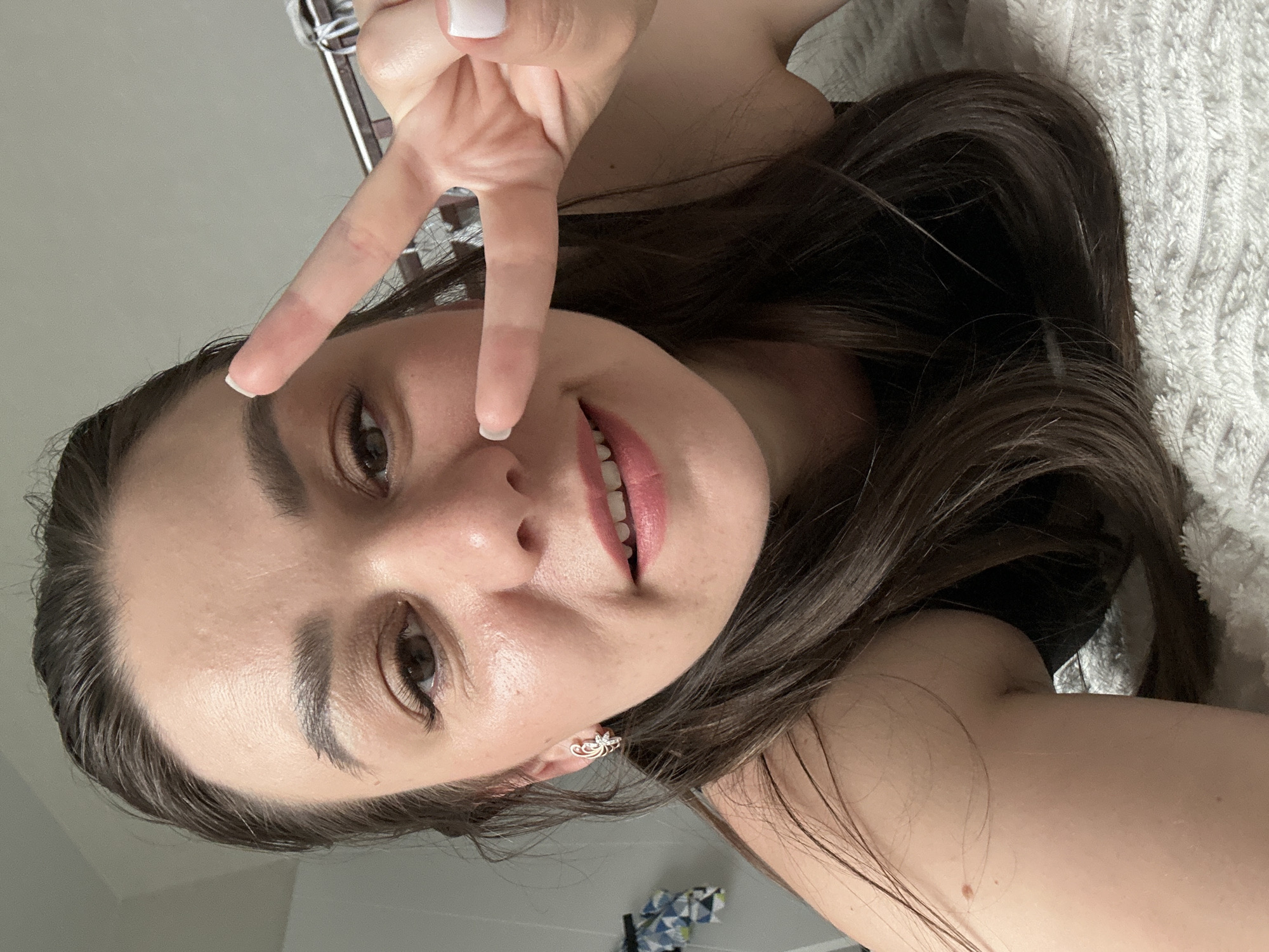 A young woman is lying down, possibly on a bed, with a white textured blanket visible in the background. She is making a peace sign with her hand. She has long brown hair and fair skin. She is wearing earrings. The scene appears to be indoors. The location in the city of Chaikovsky, Russia is unknown.