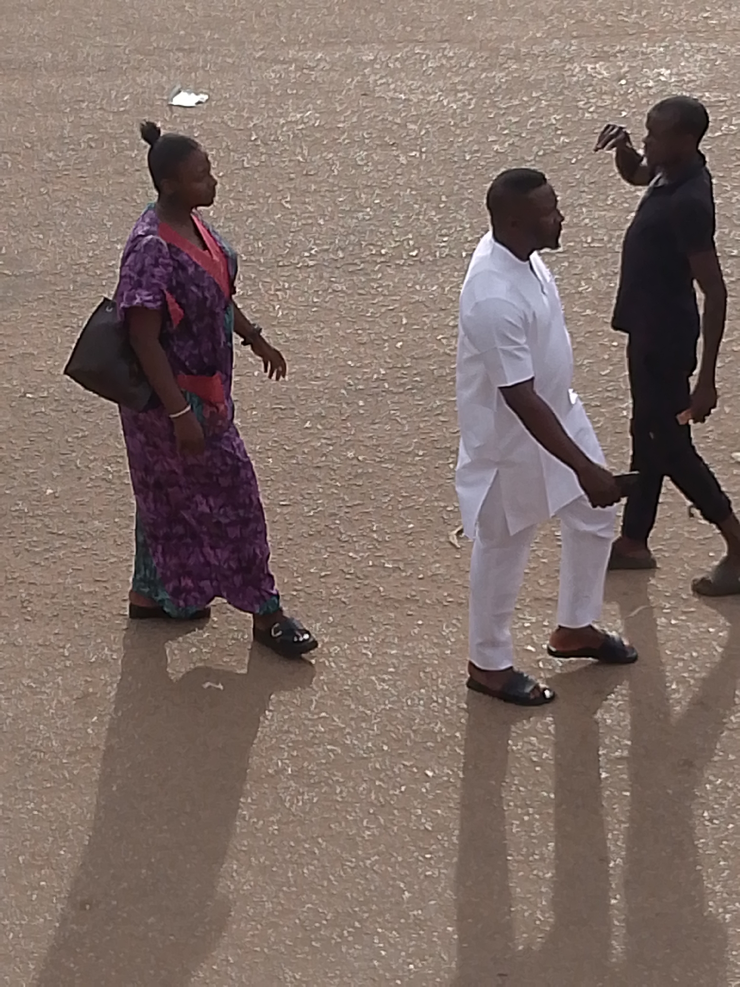 Three people are visible on a paved surface in Bukuru, Nigeria. On the left, a woman wearing a purple and pink patterned dress with a black handbag slung over her left shoulder is walking to the right. She has her hair pulled back in a bun. In the center, a man wearing a white tunic and trousers is walking to the right and looking to his left. He is holding a dark-colored object, possibly a phone, in his right hand. To the right of him and slightly behind, another man wearing a black short-sleeved shirt and dark trousers is also walking to the right. He is gesturing with his right hand towards the man in white. The ground appears to be asphalt or concrete, with reflections visible on its surface. The lighting suggests it is daytime.