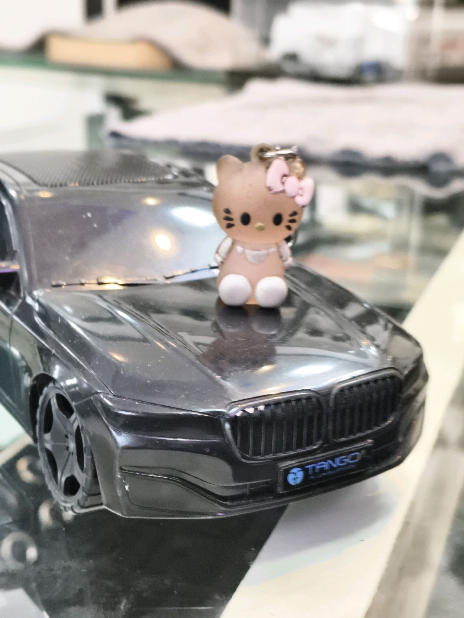 A close-up, eye-level, medium shot captures a toy black BMW car parked on a reflective surface. Perched on the hood of the car is a small, light brown Hello Kitty figurine with a pink bow. The figurine is wearing a light pink dress and has black eyes and whiskers. A silver keychain ring is attached to the top of the figurine's head. The car's grill features a license plate with a blue "TANGO" logo. The background is blurred, showing what appears to be interior elements of a store or display area with soft lighting. The overall scene suggests a display of toys or collectibles in an indoor setting. There is no indication of Pazhayangadi, India.