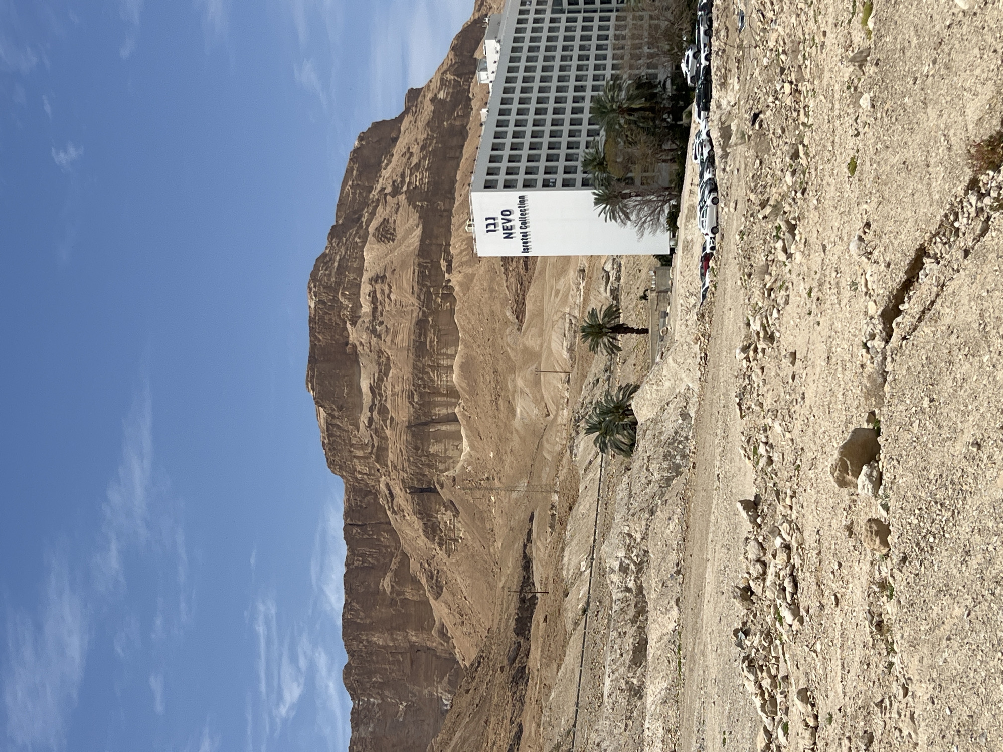 A terrestrial view features a tall, multi-story white building labeled "NEVO" with the Hebrew lettering "נבו" at the top. It's situated on a rocky, arid landscape, juxtaposed against a towering, stratified, brown mountain formation. The sky is clear blue with scattered wispy clouds. Several palm trees are scattered in front of the building. To the building's right, numerous parked cars line the edge of a rugged, unpaved area. The scene is likely in the Tamar Regional Council, Israel, based on the building signage.