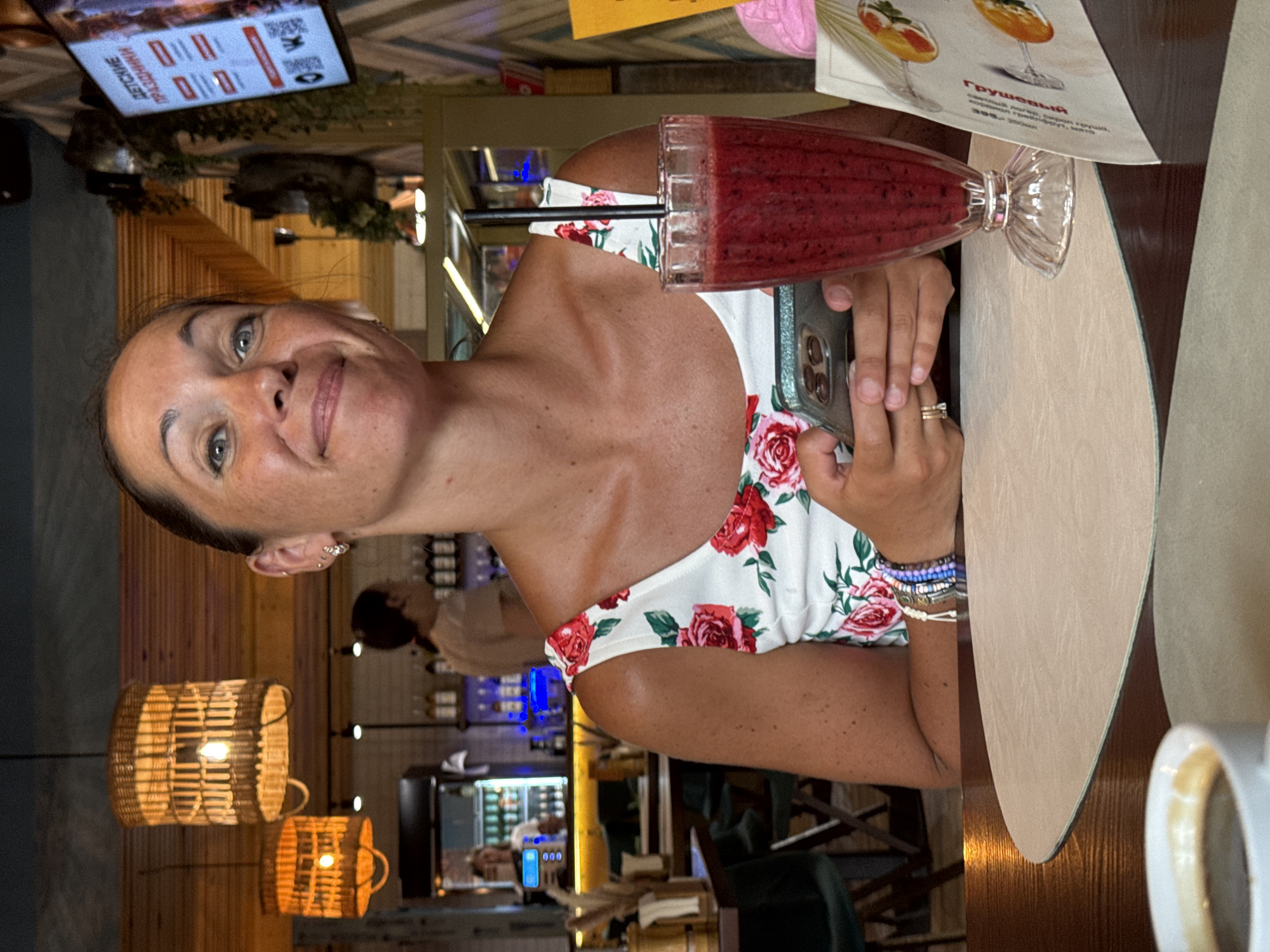 A woman in a floral-print dress holds a smartphone and a glass of berry smoothie at a table in a cafe. The cafe has wooden accents and a dark ceiling. A menu is partially visible on the right side of the table. The woman has a bright expression and is looking towards the camera. The scene appears to be in Sochi, Russia, given the Cyrillic text visible on a screen in the background that reads "ДЕТСКИЕ ПРАЗДНИКИ" (Children's Parties).