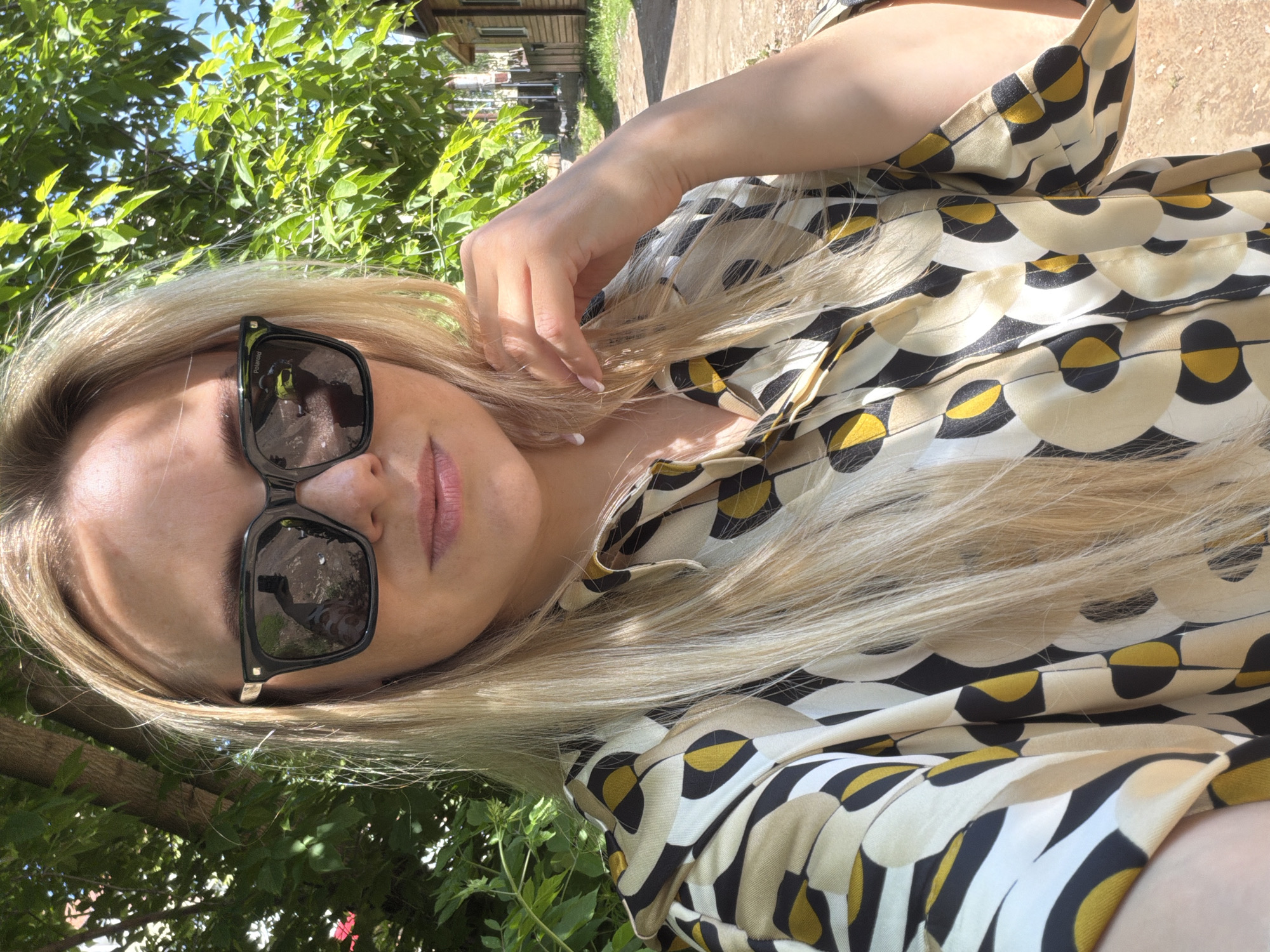 A blonde woman is depicted in a close-up selfie outdoors in the city of Votkinsk, Russia. She is wearing square-framed dark sunglasses, with a visible "Polaroid" inscription on the left lens frame. Her long, straight blonde hair falls over her shoulders. She wears a button-up shirt featuring a geometric pattern composed of concentric circles and teardrop shapes in shades of beige, white, black, and mustard yellow. Her left hand, with light-tipped fingernails, is positioned near her chin and neck. The reflection in her sunglasses shows the outline of a person holding a phone, indicating a selfie. The background features dense green foliage and tree trunks on the left. To the right, a dirt path extends into the distance, bordered by multiple wooden houses with dark roofs under a clear blue sky. Sunlight illuminates the scene, casting shadows on her forehead and highlighting the leaves.