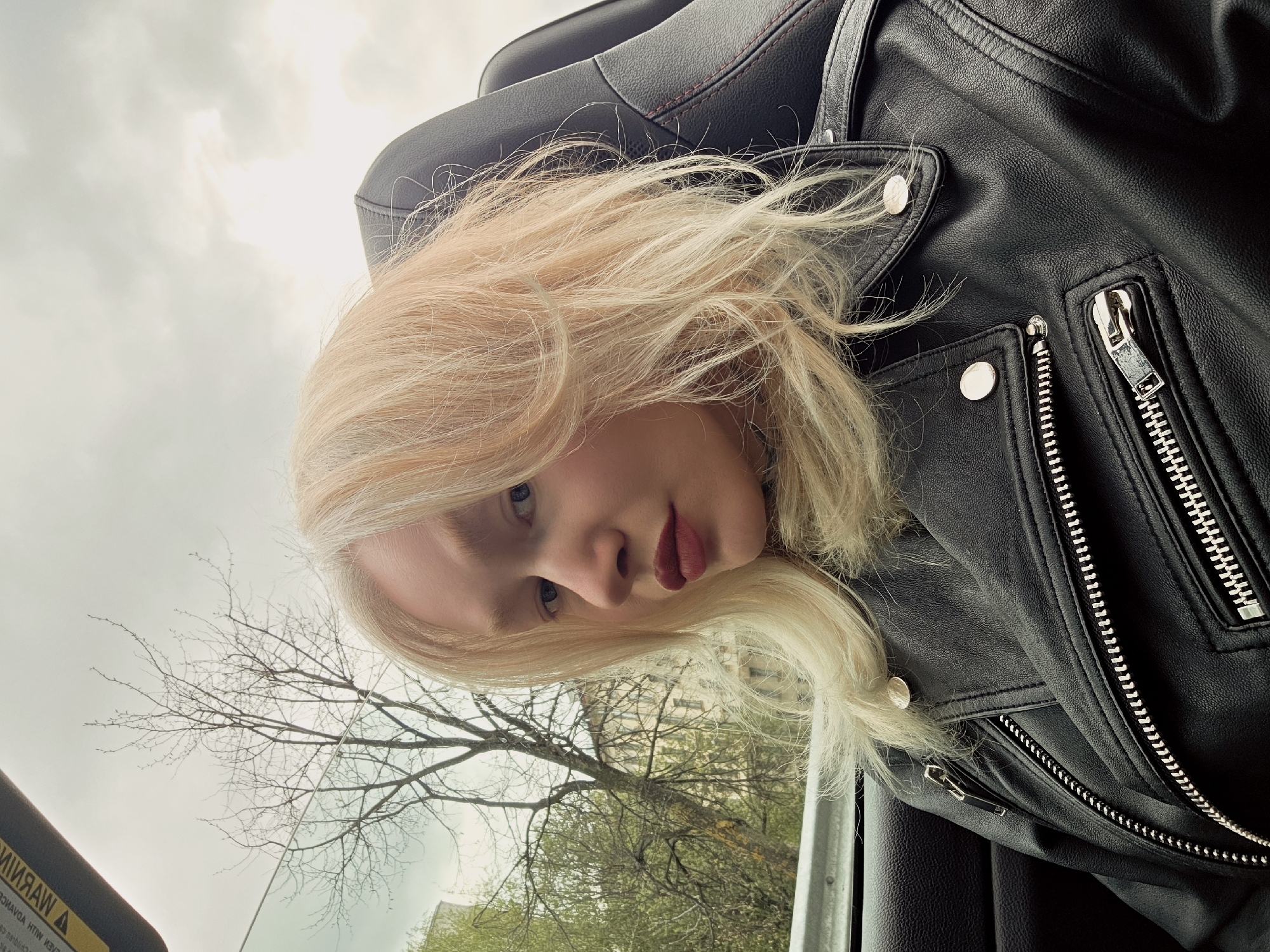 A low-angle, medium shot depicts a person with fair skin and light blonde hair inside what appears to be a car. The person is wearing a black leather jacket with silver zippers and snaps. Part of a car seat is visible behind them. Outside the car window, a tree with bare branches is seen against a cloudy sky. The lower part of the frame shows green foliage and possibly buildings in the distance.
The environment is assumed to be Saint Petersburg, Russia.