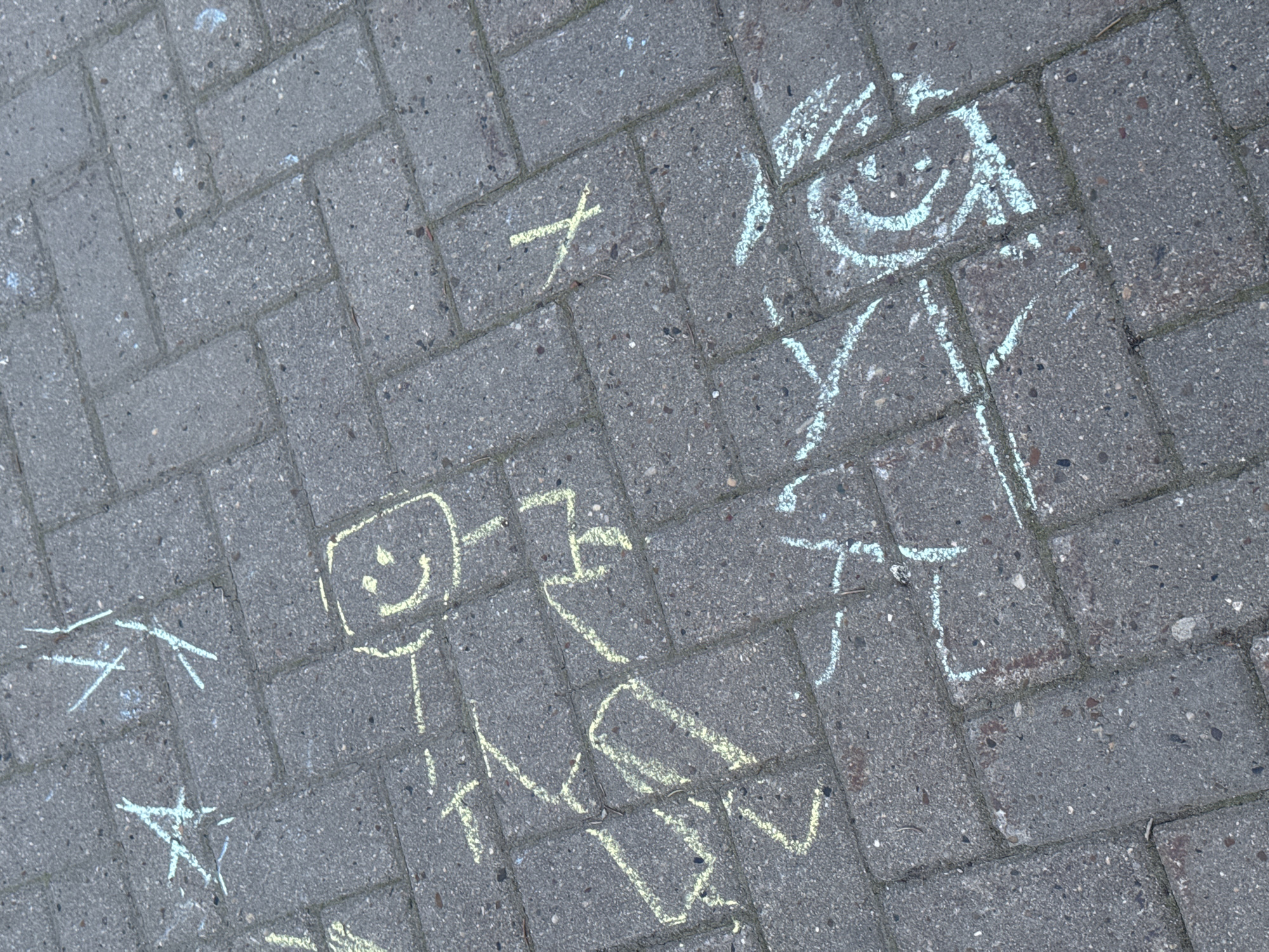 The image depicts a section of a brick-paved surface. Chalk drawings of figures and symbols are present on the brickwork. The figures include rudimentary depictions of people or characters with simple facial features. Additional geometric shapes and lines are also visible in chalk. No people, subjects, actions or interactions are physically present in the frame; only the chalk drawings on the pavement. The environment is limited to the brick paved surface. The city of Izhevsk, Russia, is not discernible from the provided image.