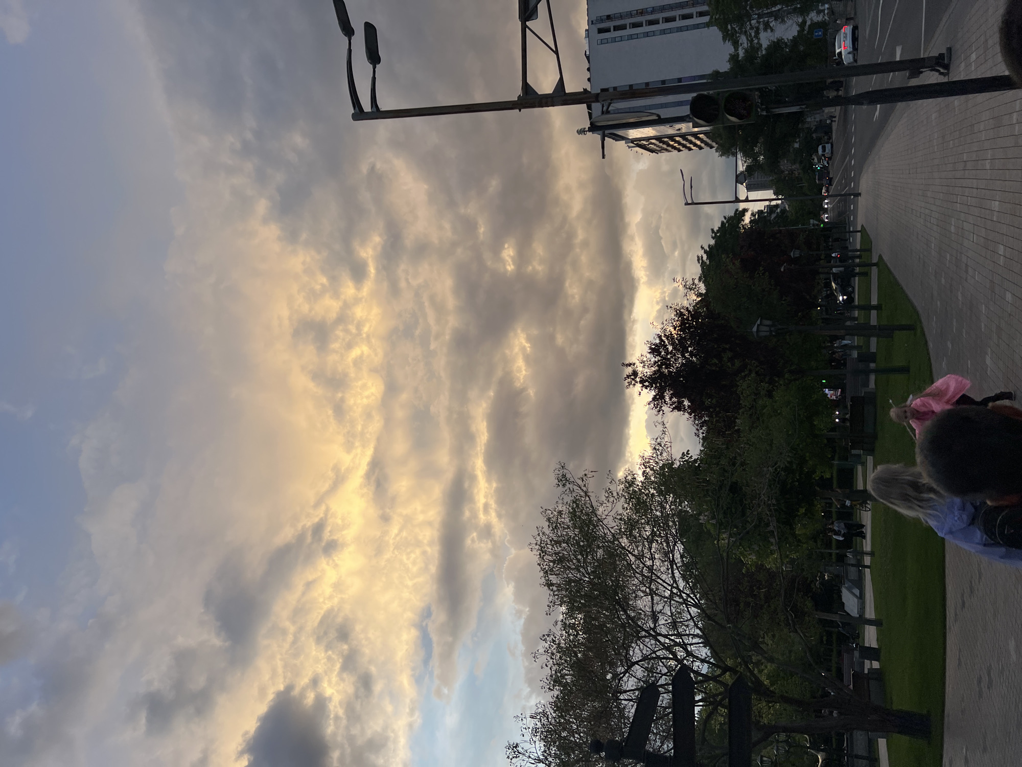 The image shows an outdoor scene in Yerevan, Armenia, likely a park or pedestrian area alongside a city street. The view is angled upwards towards a dramatic sky filled with billowing, golden-hued clouds, indicating either sunrise or sunset. The sky occupies a large portion of the frame.
In the foreground, the edge of a paved walkway or sidewalk is visible, with grass and trees lining it. Some blurred figures of people are present on the walkway. Streetlights and overhead power lines are prominent on the left side of the image, extending into the sky.
In the background, there's a glimpse of a modern high-rise building partially obscured by trees. A city street runs parallel to the walkway, with some visible traffic.