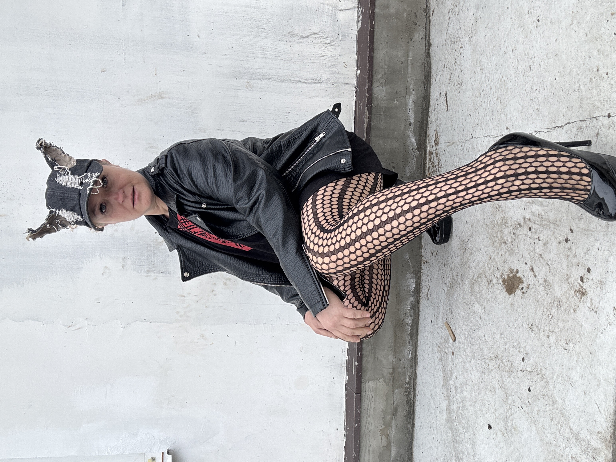 A person wearing a black leather jacket, a cap with added ear-like appendages, and fishnet stockings is posed on a concrete surface. The person is also wearing black high heels. The location appears to be outdoors near a concrete wall. Based on additional information provided, the location is Chaikovsky, Russia.
