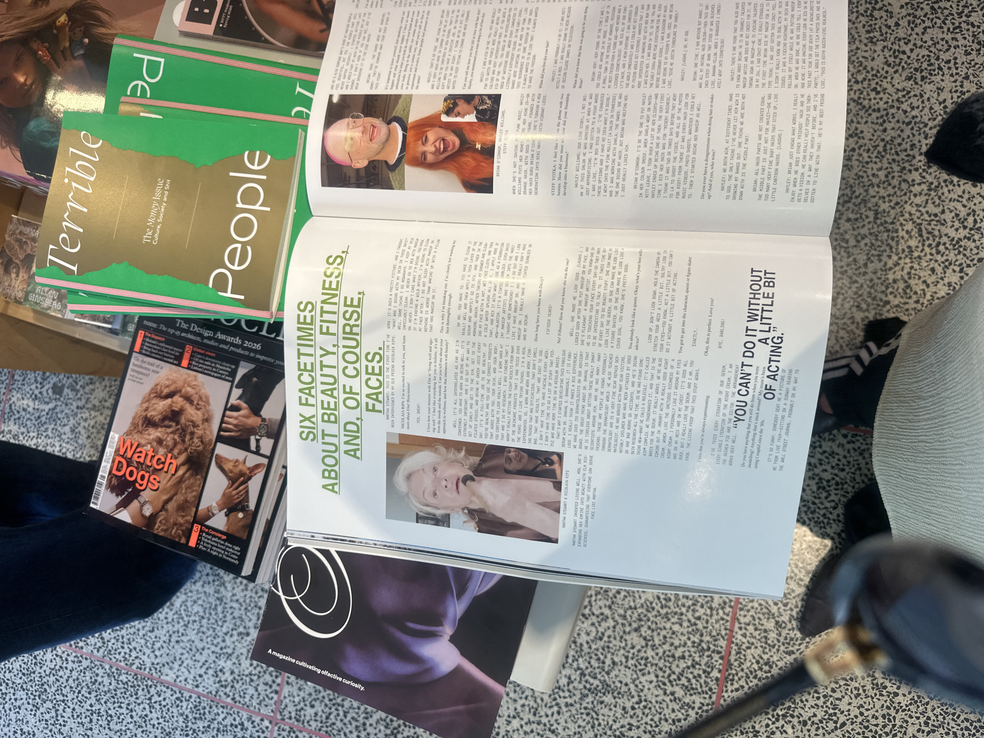 The image shows a collection of magazines and a newspaper lying on a patterned floor that appears to be indoors. The magazines include a stack of "People" magazines, with the top one titled "Terrible People" and subtitled "The Money Issue Culture, Society and Shit". Next to the stack is an open magazine, displaying an article titled "Six Facetimes About Beauty, Fitness, And of Course, Faces". This article features photographs of individuals. Other magazines in the scene are "Watch Dogs" and one with a purple cover with a white circular design. A portion of a person's leg wearing jeans is visible, along with a golf club. The background suggests an urban environment, but no specific London location is directly identifiable within the scene.