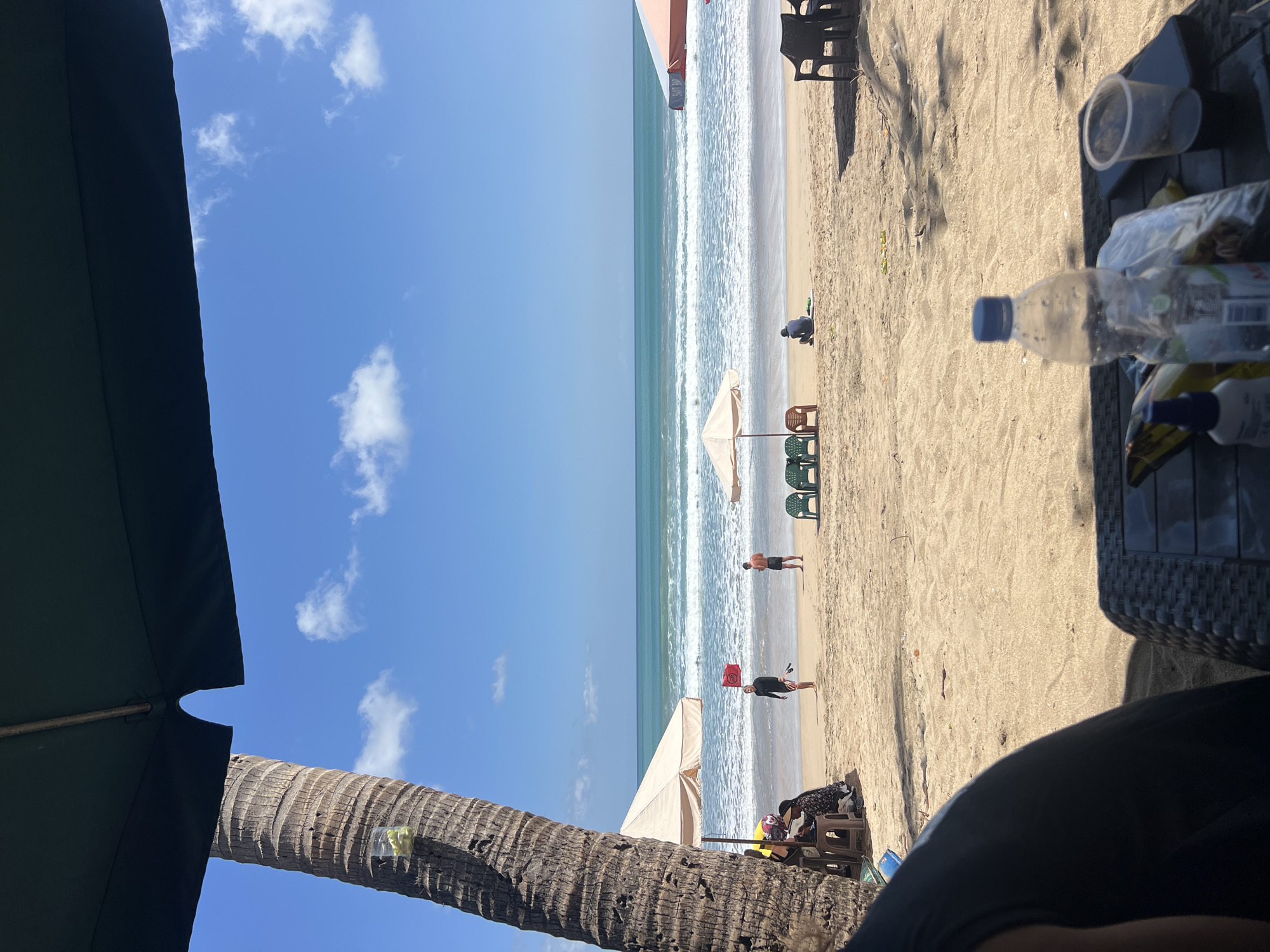 A bright, sunny day at Kuta Beach, Indonesia. The foreground is dominated by the shadow of a large umbrella and a person's arm and shoulder in a dark t-shirt, seated at a woven table. On the table are a half-full plastic water bottle, a clear plastic cup with dark liquid, a phone, and some bagged items. The middle ground features a sandy beach with several beach umbrellas, lounge chairs, and a few people scattered about. A tall palm tree is prominent on the left side of the frame, with a plastic cup attached to its trunk containing some green items. The ocean is visible in the background, with gentle waves rolling onto the shore. The sky is a clear blue with scattered white clouds. One person in the mid-ground is wearing a black wetsuit and holding a surfboard and a red flag.