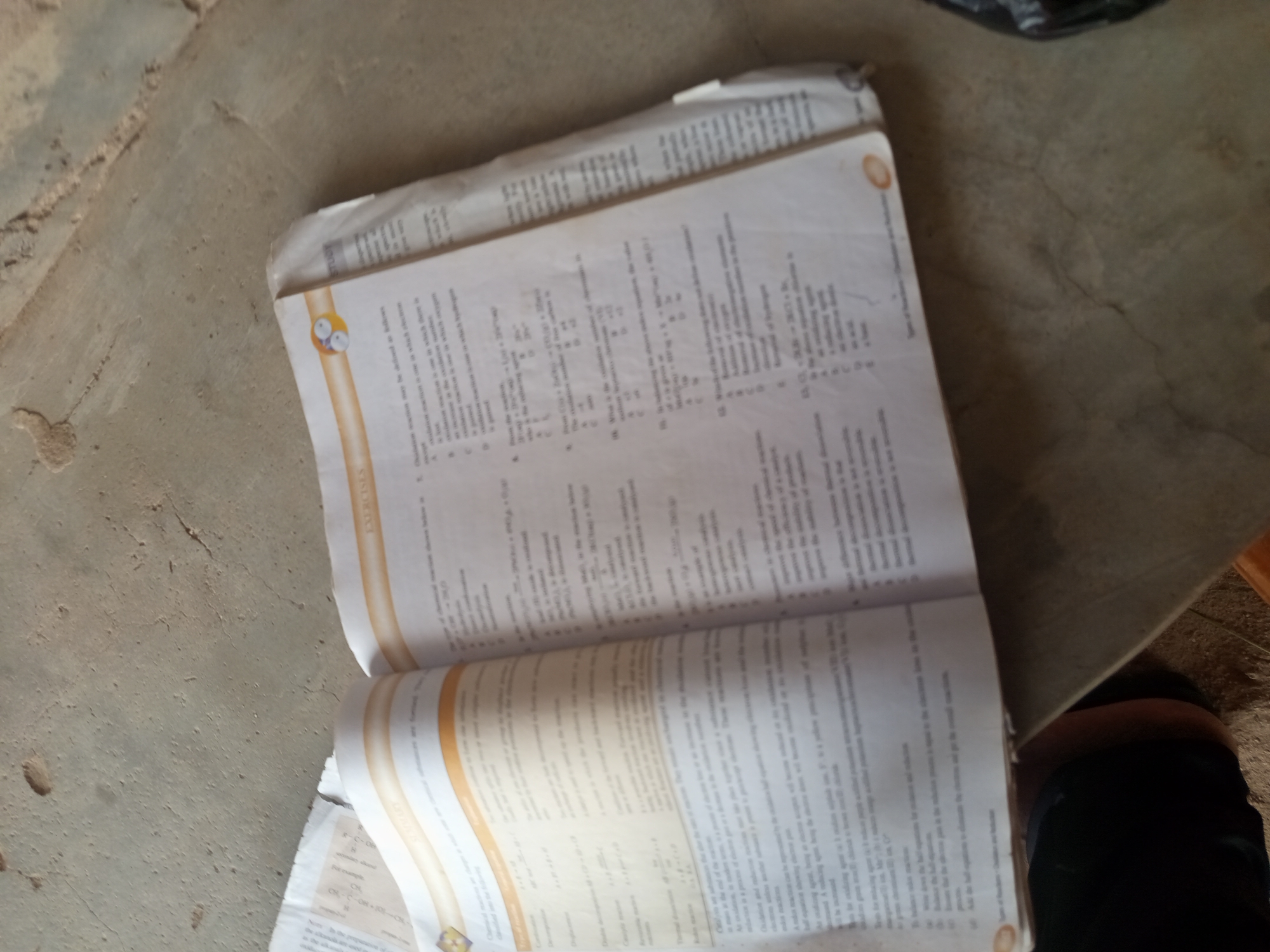 An open book lies on a textured, concrete surface. The book is open to pages with printed text and equations. The binding of the book is visible, and the pages appear to be slightly creased and worn. The concrete surface has a rough, uneven texture with visible cracks and imperfections. The lighting suggests an indoor or shaded outdoor environment. There is no discernible information about the city of Jalingo, Nigeria, or any specific people, actions, or interactions in the image.
