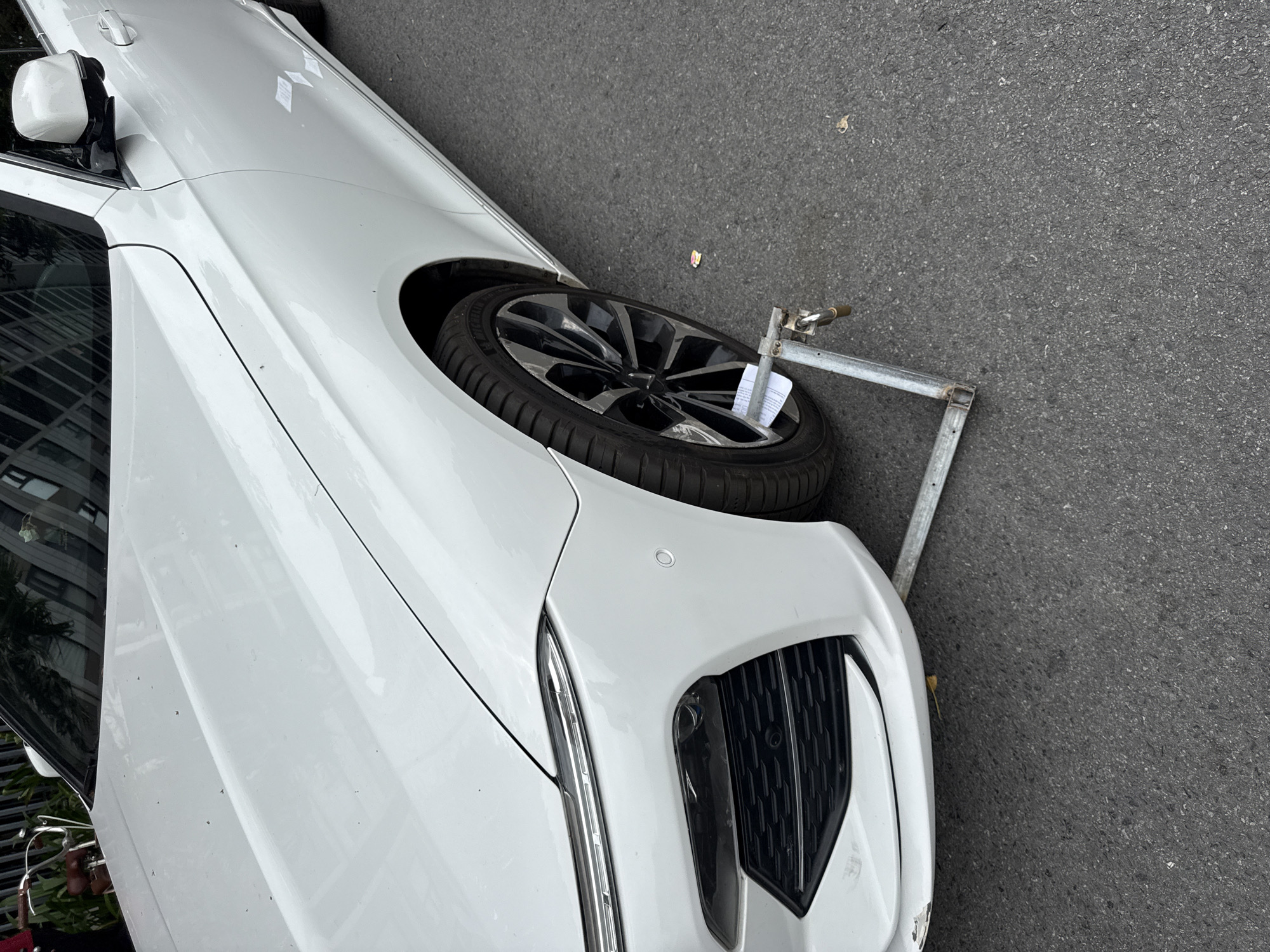 A white sedan is shown from a front-quarter perspective, parked on an asphalt surface in Hà Nội, Vietnam. The vehicle's front right wheel is immobilized by a metallic, triangular-shaped wheel clamp secured with a brass-colored padlock. A folded white paper document is inserted into the spokes of the dark, multi-spoke alloy wheel, which is equipped with a Michelin tire. The car's front bumper features a black lower grille, a central radar sensor, and a white parking sensor on the passenger side. Scuff marks and paint loss are visible on the lower left portion of the front bumper. The white bodywork shows light dust accumulation. Reflections in the vehicle's surfaces indicate nearby buildings with window patterns and greenery. The top-left corner reveals a portion of a bicycle.