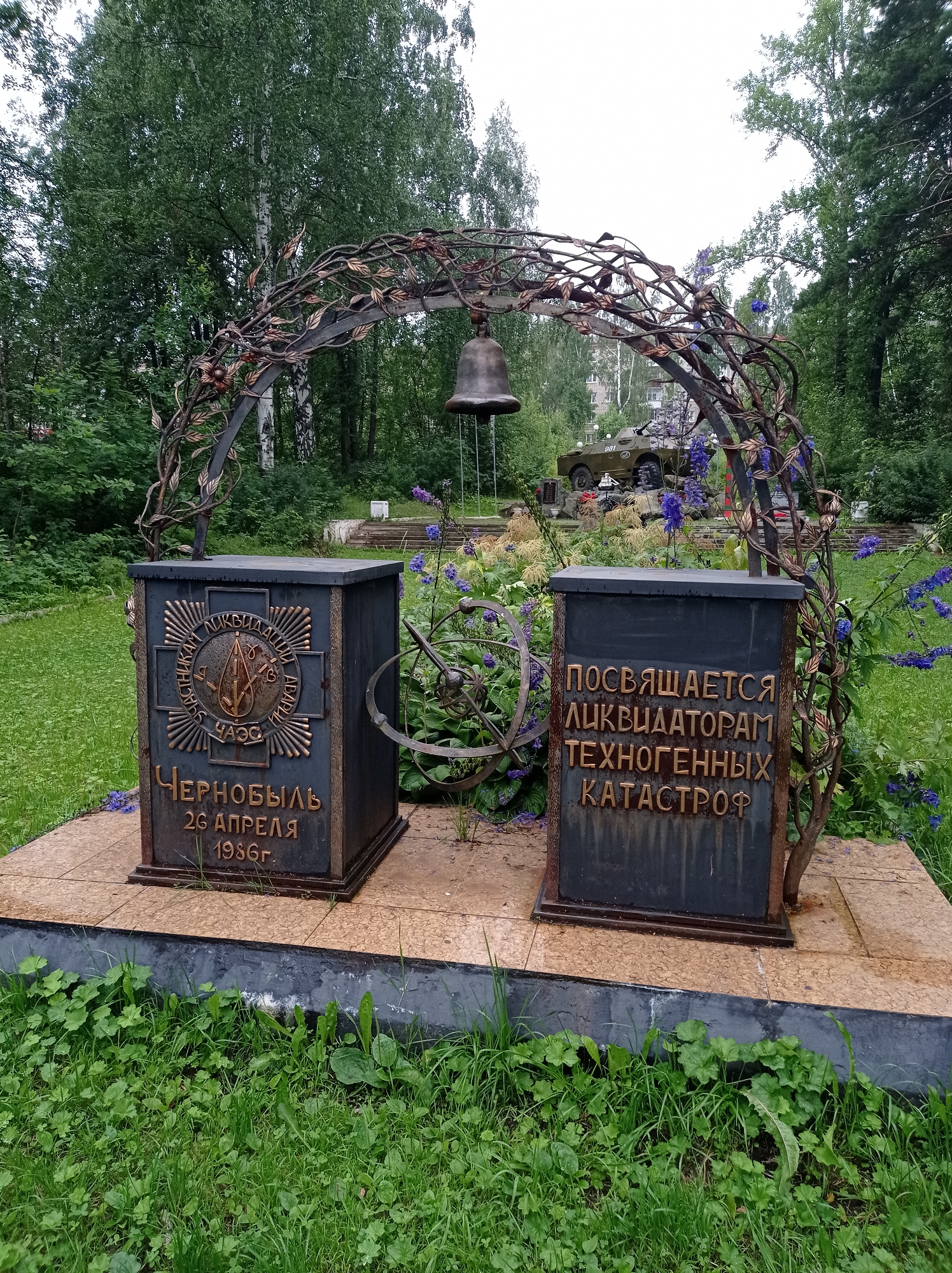 The photograph captures a memorial in the city of Degtyarsk, Russia, dedicated to the liquidators of technological disasters. The memorial consists of two dark, rectangular pedestals placed on a tiled platform, framed by a decorative metal arch resembling branches and leaves. A bell hangs from the apex of the arch.
The left pedestal features a circular emblem with radial lines and an image resembling a stylized drop within a sun-like structure. Below the emblem, large gold-colored Cyrillic letters spell out "ЧЕРНОБЫЛЬ 26 АПРЕЛЯ 1986г.", commemorating the Chernobyl disaster.
The right pedestal bears a more extensive inscription in gold-colored Cyrillic letters: "ПОСВЯЩАЕТСЯ ЛИКВИДАТОРАМ ТЕХНОГЕННЫХ КАТАСТРОФ", which translates to "Dedicated to the liquidators of man-made disasters."
Behind the memorial, a grassy area with scattered foliage and purple flowers is visible. In the background, a military vehicle, possibly an armored personnel carrier, is partially visible, along with trees and residential buildings. The environment appears to be an outdoor park or memorial site. There are no people visible in the image.