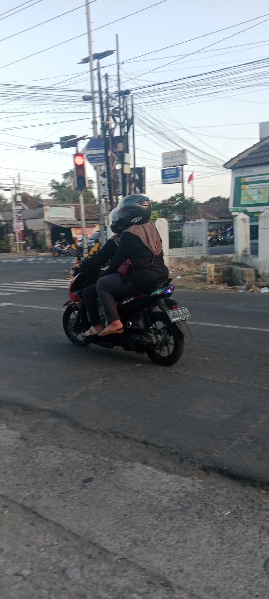 The image shows two people riding a motorcycle on a street in Maguwoharjo, Indonesia. The motorcycle is black and red, with a license plate that reads "AB 3028 RB". The rider and passenger are both wearing helmets. The rider is wearing a dark-colored jacket and pants, and the passenger is wearing a black hijab and a brown scarf. They are riding on a paved road with a pedestrian crossing visible in the foreground. In the background, there are buildings, power lines, and traffic lights. The sky is clear and the sun is setting.