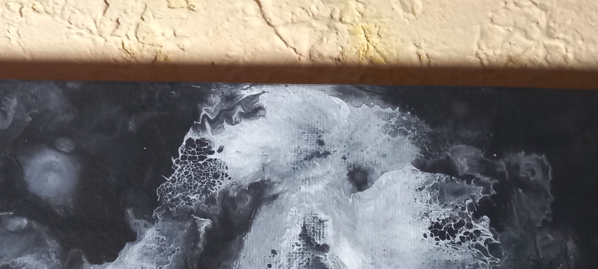 The media file presents a detailed view of an abstract painting, likely a fluid art or acrylic pour piece, set against a textured background. The artwork predominantly features a monochrome palette of black, white, and shades of grey. It showcases dynamic, organic patterns with flowing, marbled sections alongside areas exhibiting intricate cellular or lacing textures, particularly where lighter tones emerge from or blend with darker ones.
Above the abstract artwork, occupying roughly the top quarter of the image, is a highly textured, light beige or pale yellow surface, possibly a stucco wall or ceiling. This surface displays an uneven, rough quality with visible ridges and depressions. A prominent, dark, horizontal shadow is cast by this textured surface directly onto the top edge of the abstract painting, indicating a light source from above or the side that highlights the texture of the wall.
The scene is static, devoid of any visible people or ongoing activities. The focus is entirely on the visual textures and patterns of the abstract art and its immediate architectural setting. No text is visible within the image. The time of day is not discernible, and there are no specific location cues that would identify it as Kryvyi Rih, Ukraine.