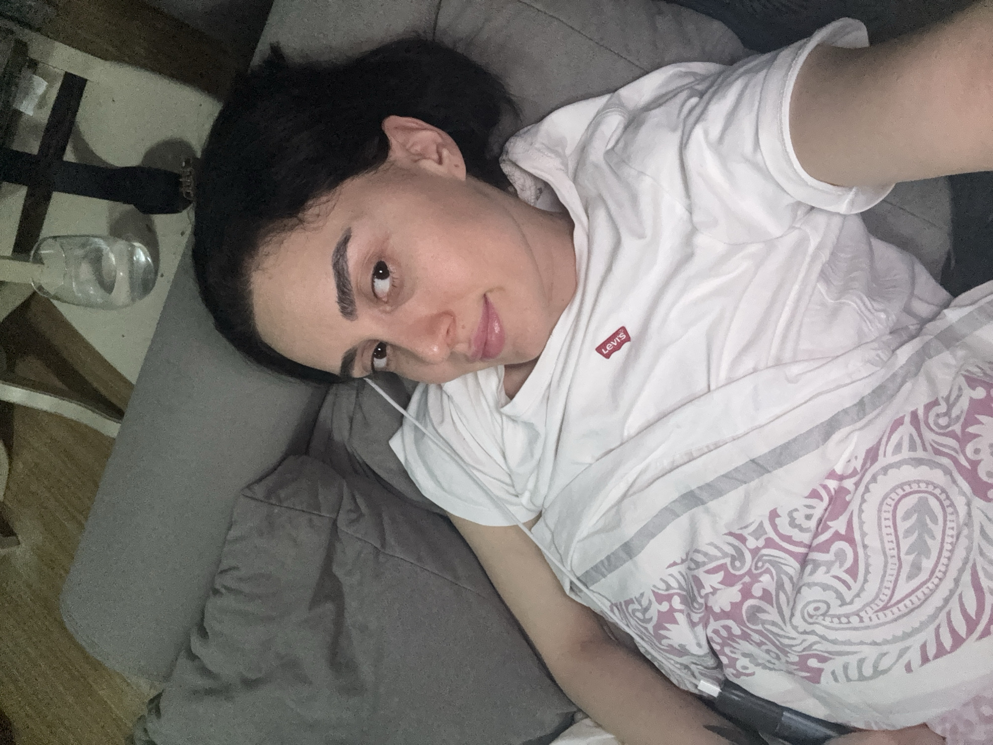 A woman is lying on a gray couch in an indoor setting. She has dark hair and is wearing a white t-shirt with a red Levi's logo. A white and pink patterned blanket covers her lower body. A glass of water sits on a table next to the couch. The location appears to be in a residential setting, but there's not enough information to specify whether it's in Tbilisi, Georgia.