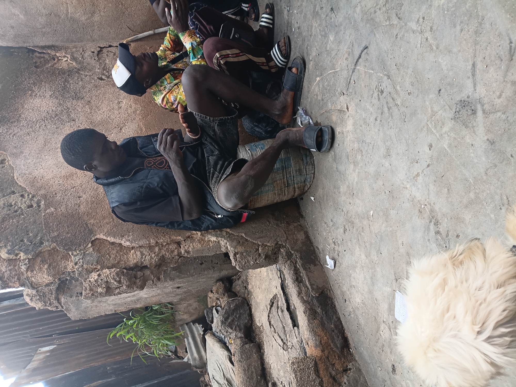 The image depicts a scene in Gwong, Nigeria. Three individuals are visible, with one person in the foreground sitting and looking down at their hands. This person is wearing a black jacket and dark shorts, and has a bracelet on their wrist. To their right, another person is seated, wearing a colorful patterned shirt and dark pants. They are also wearing sandals. A third person, mostly obscured, is seated further back. The setting appears to be an outdoor area with a rough, textured wall on the left and a concrete floor. Some corrugated metal roofing is visible at the bottom left, and greenery is growing from a gap in the structure. The overall lighting suggests it might be daytime. The atmosphere seems casual, possibly a moment of rest or conversation. There is no visible text in the image.