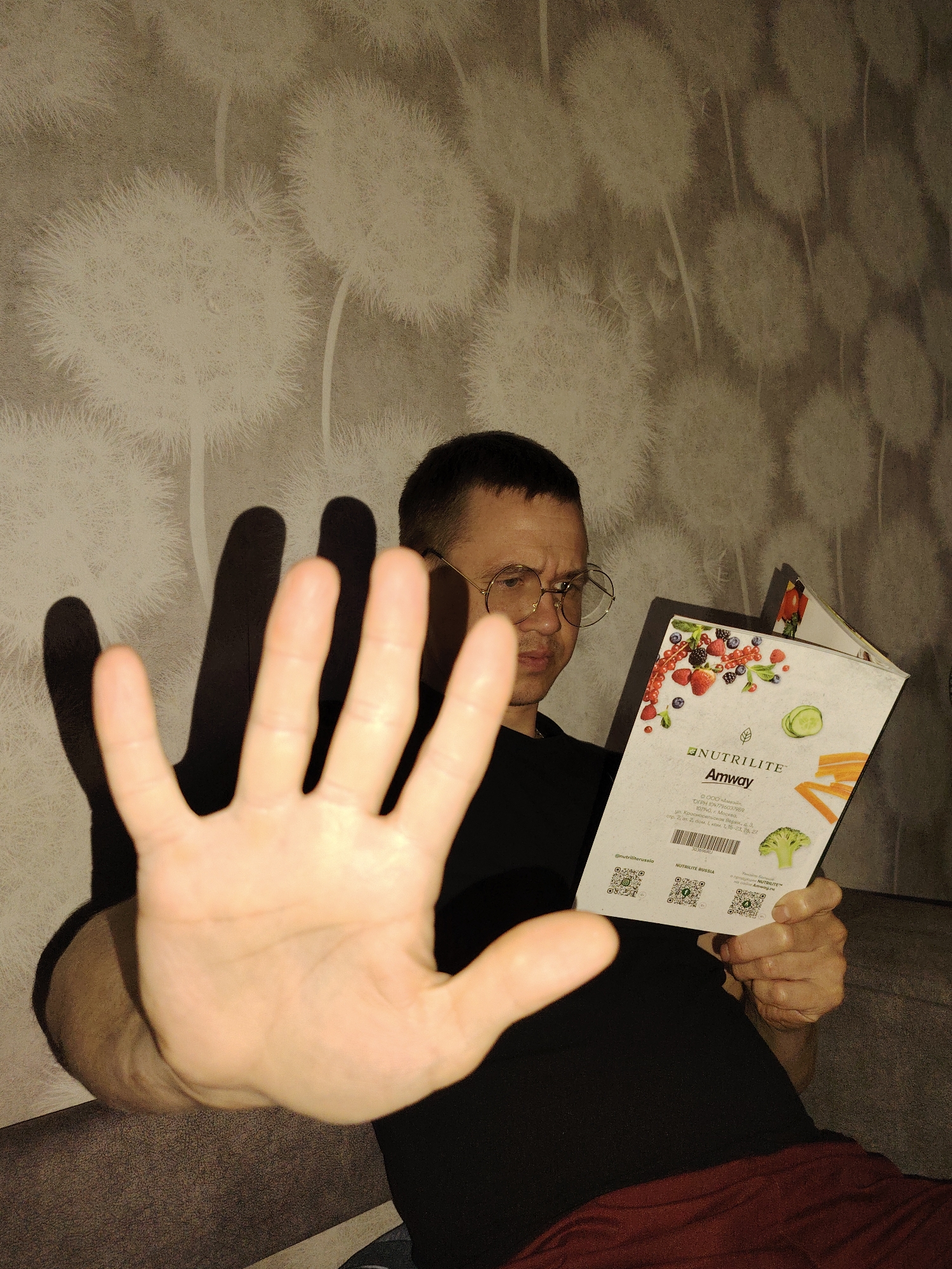 A person with short, dark hair and round, wire-frame glasses is seated and holding up their left hand, palm forward, in a stop gesture. The person is wearing a black shirt and red pants. They are holding an open catalog in their right hand, displaying the "NUTRILITE Amway" logo along with images of fruits and vegetables. The catalog lists an address in Moscow, Russia. The background features a wallpaper with a dandelion design. The environment appears to be indoors, possibly a living room. The city of Chaikovsky is not evident in the image.