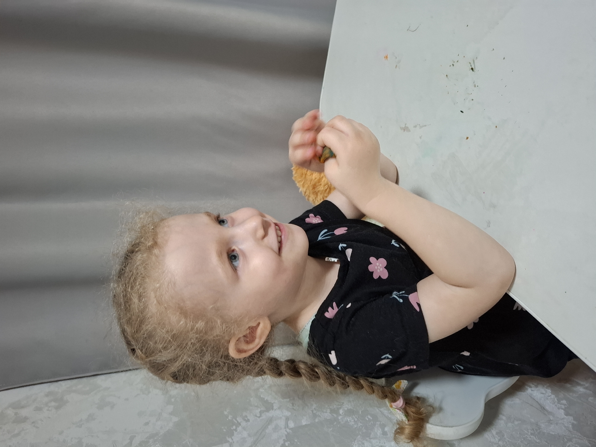 A fair-skinned girl with blonde hair braided in a single plait is positioned near the edge of a white table. She is smiling and appears to be holding a small object in her hands. She wears a black shirt adorned with a floral pattern. She is on a stool. The backdrop consists of a gray curtain and a tiled floor. The location is Chaikovsky, Russia.