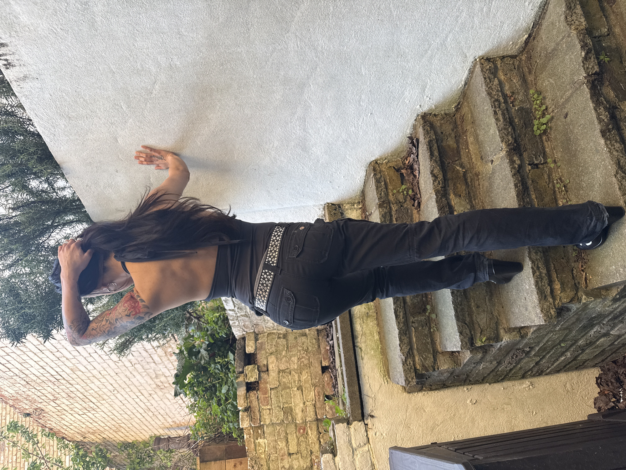 An outdoor, slightly rotated image captures a person standing on weathered stone steps amidst a rustic setting, likely in London, United Kingdom. The individual has long, dark hair and is dressed in a dark-colored top, dark jeans, and a prominent studded belt. They are also wearing dark shoes or boots. Visible tattoos adorn their left arm, featuring intricate patterns, and a smaller tattoo is on the back of their right hand. The person's back is partially exposed, revealing shoulder blades, and one hand is raised towards their head, suggesting a pose.
The scene features old, irregular stone steps that are partially covered with small green plants and dried leaves. To the left of the person is an aged brick wall, partially obscured by green ivy and dense shrubbery, possibly rosemary. To the right, a lighter-colored wall, likely concrete or stucco, appears. At the bottom right, a dark-colored bin or container is visible. The lighting is bright and diffuse, indicating daytime, possibly under an overcast sky or during a soft light period like morning or late afternoon. No specific emotions are discernible as the person's face is largely obscured. There is no visible text in the image.