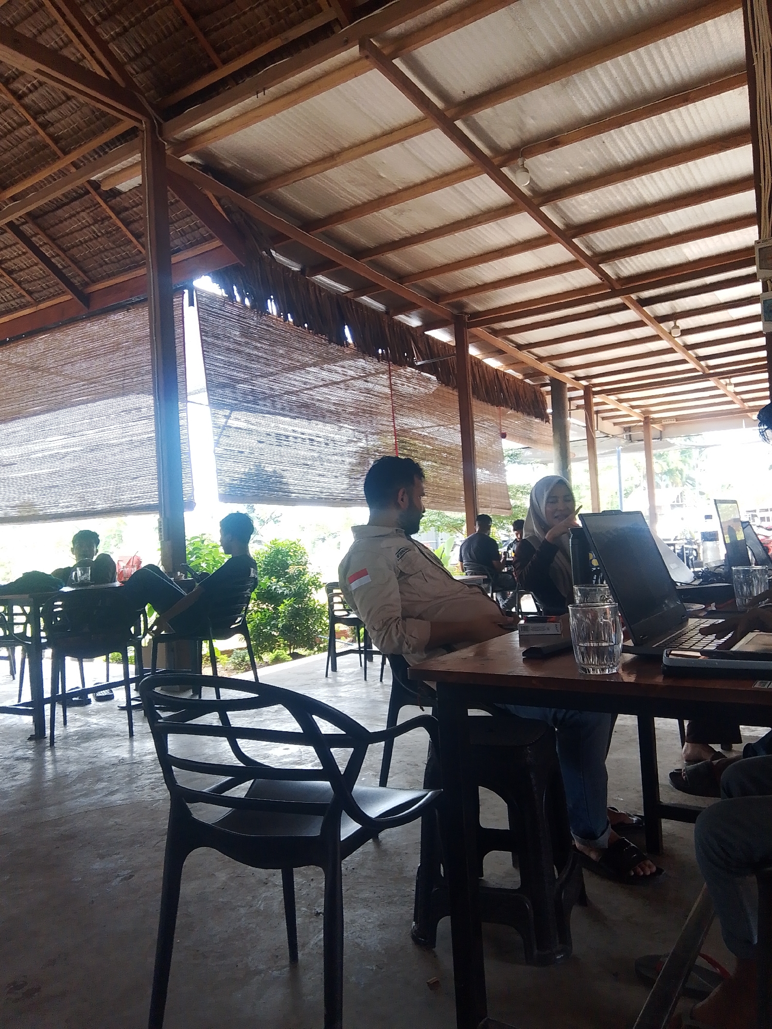 The image depicts an indoor/outdoor cafe setting with a thatched roof and open sides, suggesting a tropical or sub-tropical location, possibly in Meunasahmee, Indonesia, given the context. Several individuals are seated at tables, engaged in various activities.
In the foreground, a person wearing jeans and dark sandals is seated at a wooden table, with their legs partially obscured by the table and a black plastic chair. They appear to be interacting with a laptop, as one is visible on the table. To their right, another laptop is open, and a glass of water sits on the table. Further to the right, another person's legs, wearing dark pants and sandals, are visible.
In the mid-ground, a man in a light-colored shirt is seated at a table, facing left. He has a beard and appears to be engaged in conversation or work. A woman wearing a hijab and a black top is seated next to him, also at the table, and she appears to be gesturing with her hand. Another laptop is visible on their table. Behind them, other individuals are seated at tables, some with drinks.
The environment includes concrete flooring, wooden structural elements for the roof, and woven bamboo blinds along the open sides of the cafe. Lush green foliage is visible outside the cafe, indicating a natural setting. The lighting suggests it is daytime, with natural light filtering through the open sides and from overhead lights. The overall atmosphere appears relaxed, with patrons engaged in work or leisure.