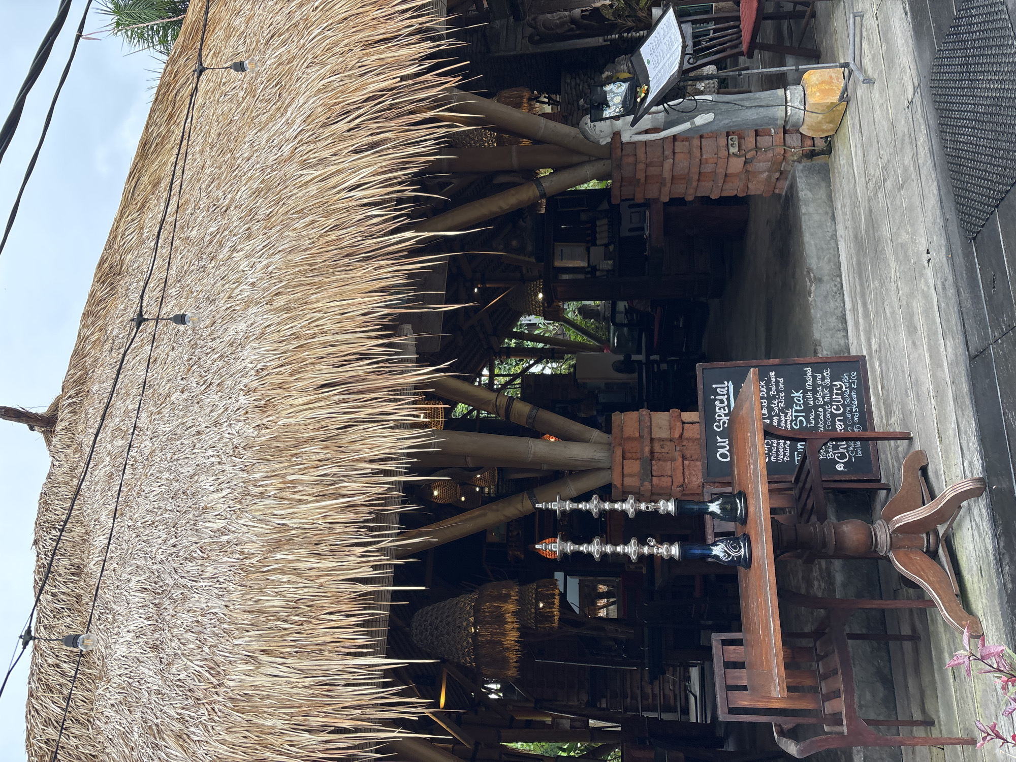The image depicts the outdoor seating area of a restaurant with a thatched roof, likely located in the Ubud District of Indonesia, given the architectural style. The thatched roof is supported by bamboo poles and has hanging lights. In the foreground, a wooden table with two chairs is set up, featuring two decorative hookah pipes. A blackboard menu stands next to the table, listing specials like "Ubud Duck," "Steak," and "Chicken Curry." A statue of a person is positioned near the menu, and a menu holder is visible behind it. The ground is made of weathered wooden planks. The background shows the interior of the restaurant with more seating, bamboo structures, and dimly lit lamps. Lush greenery is visible around the building, suggesting a tropical environment. The sky appears overcast. There are no people visible in the immediate foreground or engaged in any activities.