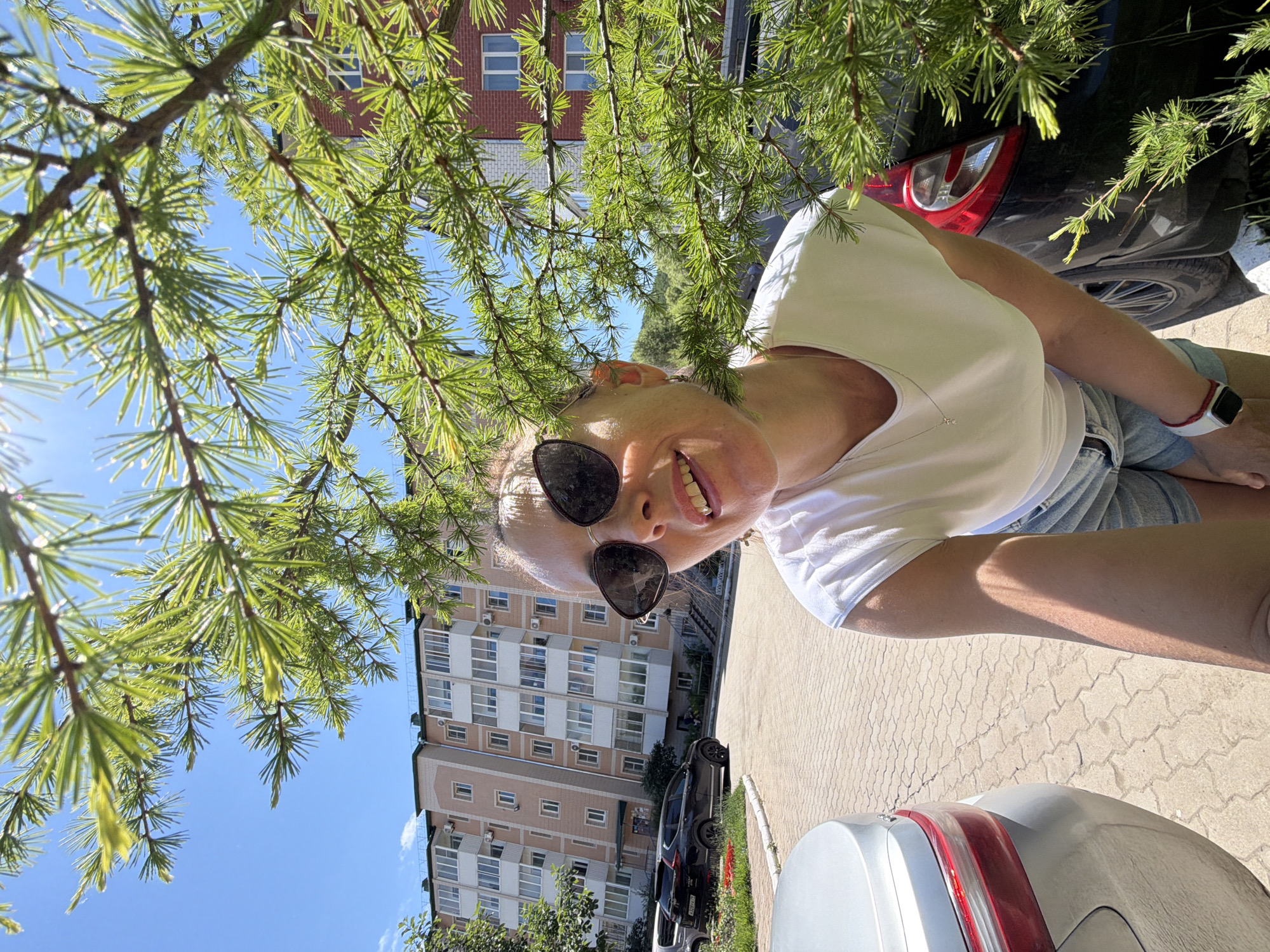 A woman is taking a selfie outdoors, partially obscured by the branches of a coniferous tree. She is wearing a white t-shirt, denim shorts, and sunglasses. Her mouth is slightly open, and she appears to be smiling. Behind her, a multi-story apartment building is visible, along with parked cars and a paved walkway. The environment suggests a residential area in Birobidzhan, Russia. The lighting indicates it is daytime, with a clear blue sky.