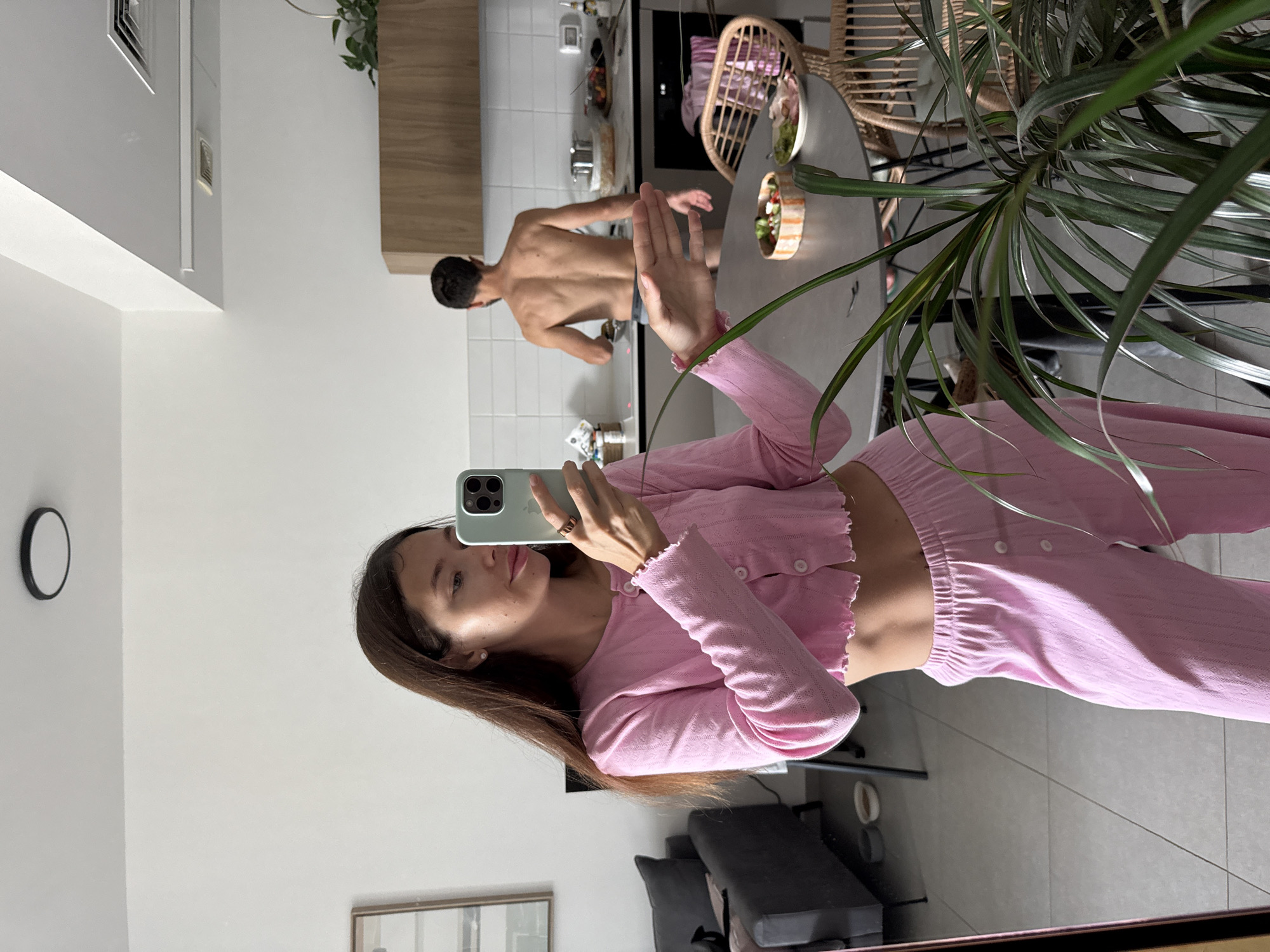 A woman is taking a selfie in a mirror. She has long brown hair and is wearing a pink outfit. A man is visible in the background, shirtless and cooking in a kitchen. The kitchen has white tiles and wooden cabinets. A dining table with a rattan chair is also visible. The presence of an Israeli electrical outlet and other kitchen elements suggest the location is likely Tel-Aviv, Israel. A clock is on the wall. A plant is in the right foreground.