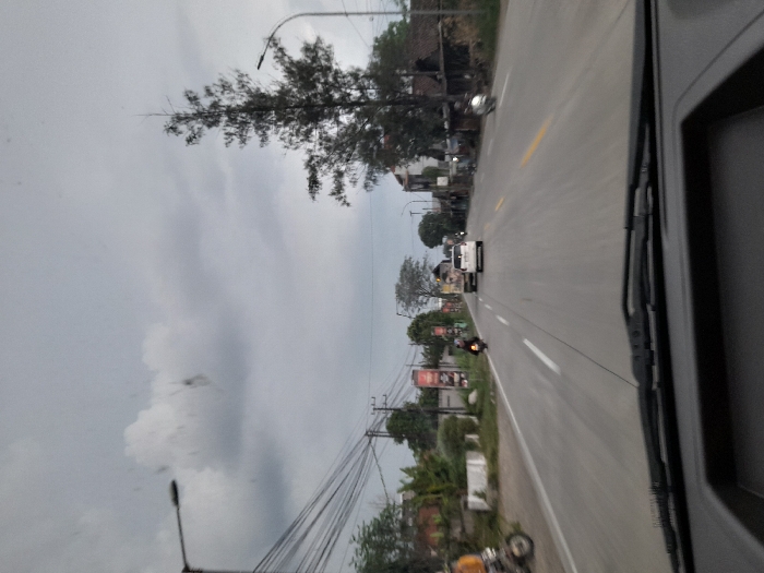 The image presents a forward-facing view from inside a moving vehicle on a two-lane asphalt road in Temuwulan, Indonesia. The sky above is entirely overcast with a uniform layer of grey clouds. The road features distinct white painted lane markings and solid shoulder lines, with yellow central lines visible further in the distance.
On the left side of the roadway, a network of overhead utility cables is visible, supported by poles. Several commercial signs, predominantly red and white, are affixed to structures along this side. A motorcycle with a rider, largely obscured, is in the immediate foreground on the left, followed by another motorcycle with a rider wearing dark clothing, traveling away from the camera.
On the right side of the road, a tall, slender coniferous tree stands prominently. Traffic in the opposing direction includes a light-colored SUV or minivan, followed by a larger utility truck, both moving away from the viewer in the right lane. A third motorcycle, with a rider wearing a light-colored helmet, is observed approaching in the far-right lane. Streetlights are mounted on poles on both sides of the thoroughfare. Various buildings, including residential and commercial structures, line both sides of the road, indicating a populated area. No explicit interactions beyond general vehicular traffic flow are discernible.