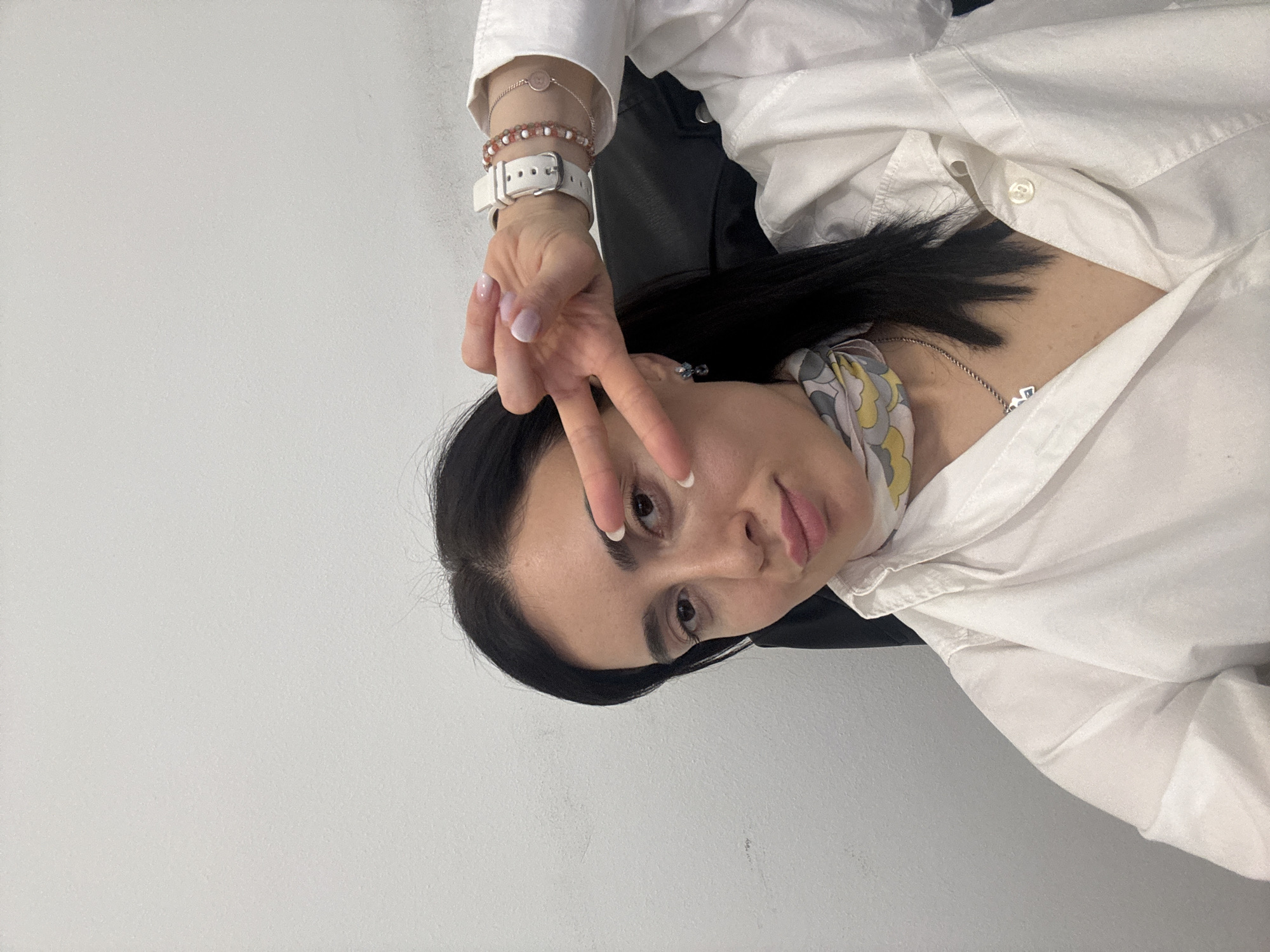 A woman with dark hair and fair skin, wearing a white button-down shirt, sits in a dark chair against a plain, light gray wall. She is making a peace sign with her hand. The scene is indoors.