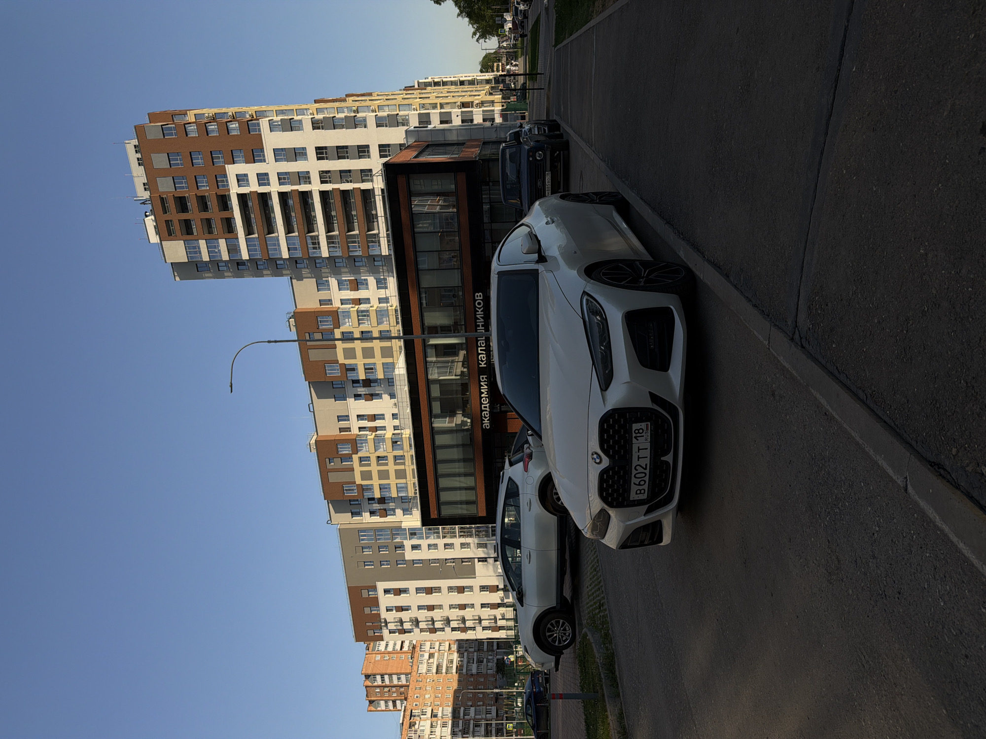 The image captures an urban scene in Izhevsk, Russia, featuring modern apartment buildings with a mix of brown, beige, and white facades. A sign with the inscription "академия калашников" ("Kalashnikov Academy") is visible on one of the buildings. Parked along the roadside are several cars, including a prominent white BMW with the license plate "B602 TT 18 RUS," and possibly a Kia Rio. The road is asphalt, and the sky is clear and blue. There are no people visible in the frame.