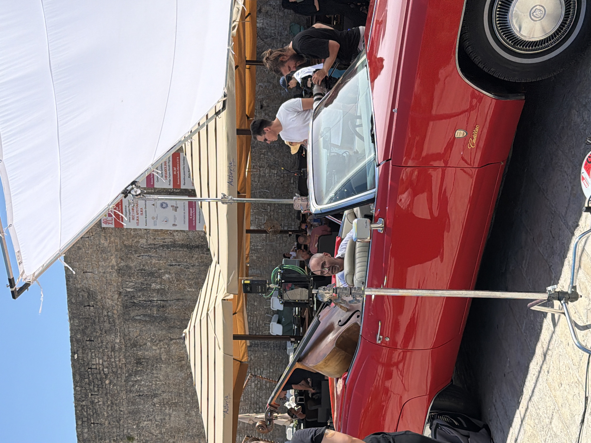 The photograph captures a scene set in Budva, Montenegro, with a prominent red Cadillac convertible as the central focus. The car is parked on a cobblestone surface, possibly a street or square, with a large, ancient stone wall forming the backdrop. Above the car, a white diffusion panel is suspended by a metal frame, suggesting a film or photo shoot is in progress.
Inside the Cadillac, a man with a bald head and a goatee is seated in the driver's seat, wearing a white shirt. To his right, a large upright bass is positioned in the passenger seat, its dark wood contrasting with the red interior of the car. In the background, several beige umbrellas provide shade, likely for an outdoor seating area, with glimpses of people seated beneath them.
To the right of the car, two men are visible. One, wearing a white t-shirt and sunglasses, appears to be observing the scene. The other, with a beard and hair tied up, is holding a professional camera with a lens, seemingly engaged in filming or photography. Behind them, a large poster is affixed to the stone wall, featuring text in a language that appears to be Serbian or Croatian, and multiple logos, possibly indicating a cultural event or festival. The sky above is a clear, bright blue, indicating a sunny day. The overall impression is that of a staged production or event taking place in a historical urban setting.