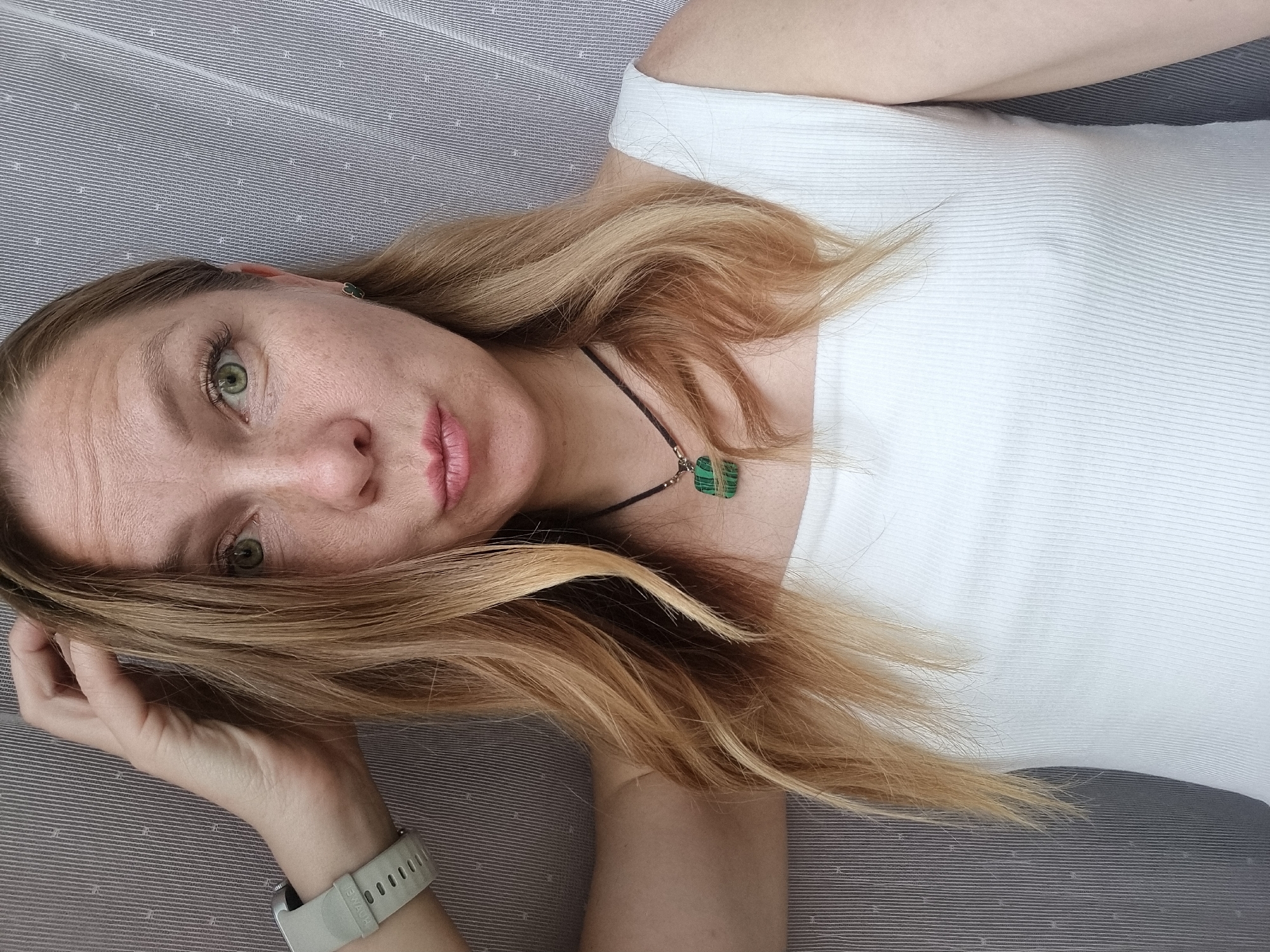 A medium shot, front-facing selfie of a fair-skinned woman with blonde hair that has blonde highlights. She is wearing a white, ribbed, sleeveless top and a dark cord necklace with a green pendant. Her left arm is bent with her hand resting on her head. She has a grey smartwatch on her left wrist. She has on a green stud earring in her right ear. The background is a grey striped curtain. The woman's lips are pursed. The image is not in Chaikovsky, Russia.