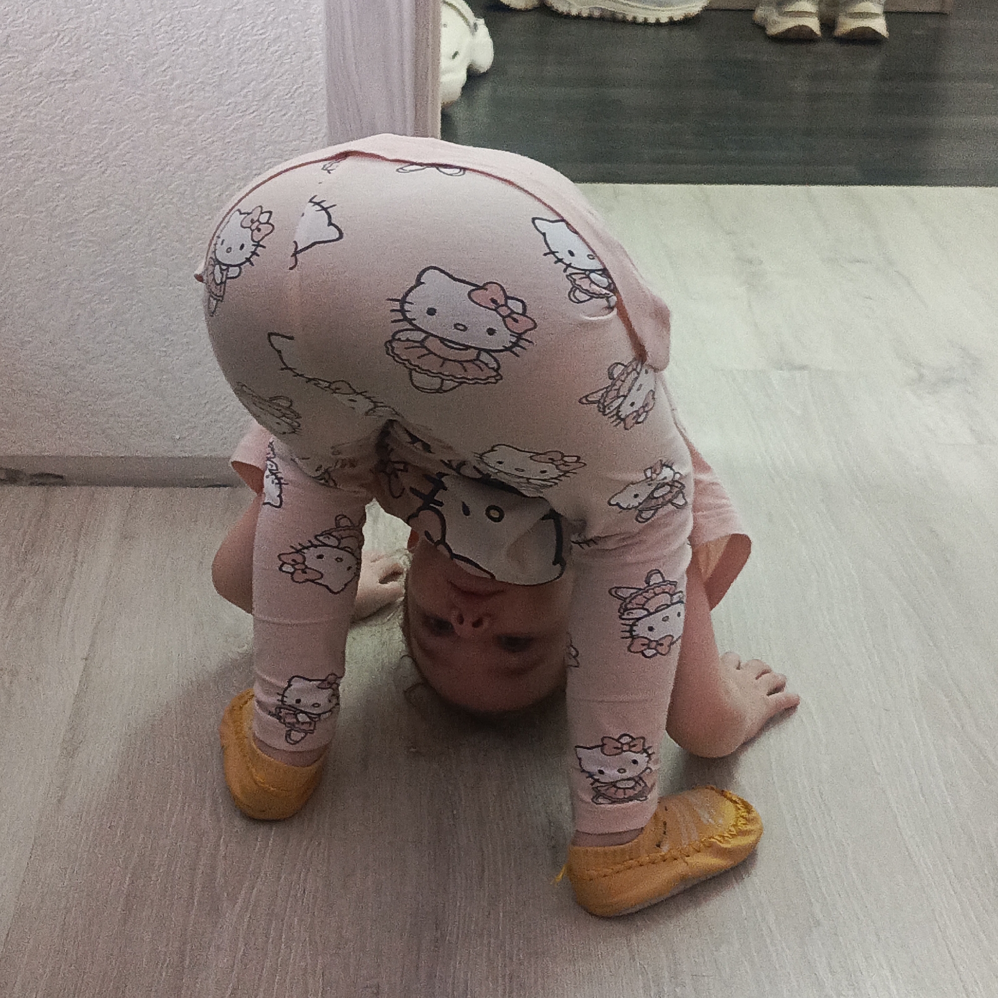 A young child, wearing pink pajamas adorned with "Hello Kitty" patterns and yellow shoes, is bent over with their head near the floor. The child's face is visible upside down. The child appears to be in a room with light-colored wood-like flooring and light-colored walls. The location is in the city of Марковский, Russia. Other shoes are visible in the background.