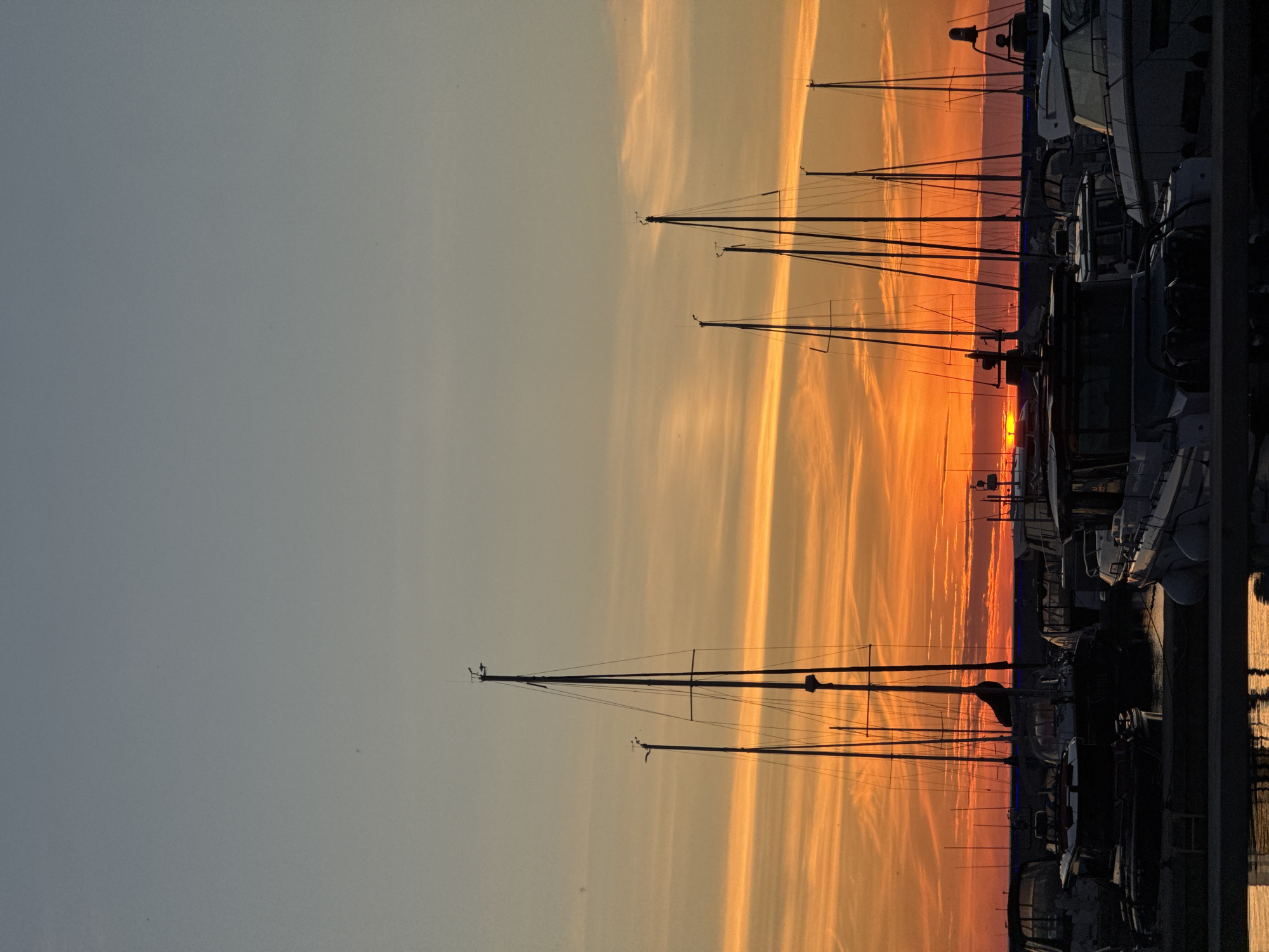 The image depicts a marina in Tallinn, Estonia, at sunset. Silhouetted sailboats and motorboats are docked in the foreground, with their masts rising towards the sky. The sky is a vibrant gradient of orange and yellow from the setting sun, with thin, wispy clouds stretching across. The water in the foreground reflects the colors of the sky and the silhouettes of the boats. No people are visible in the image. The overall atmosphere is serene and tranquil, characteristic of a harbor at dusk.
