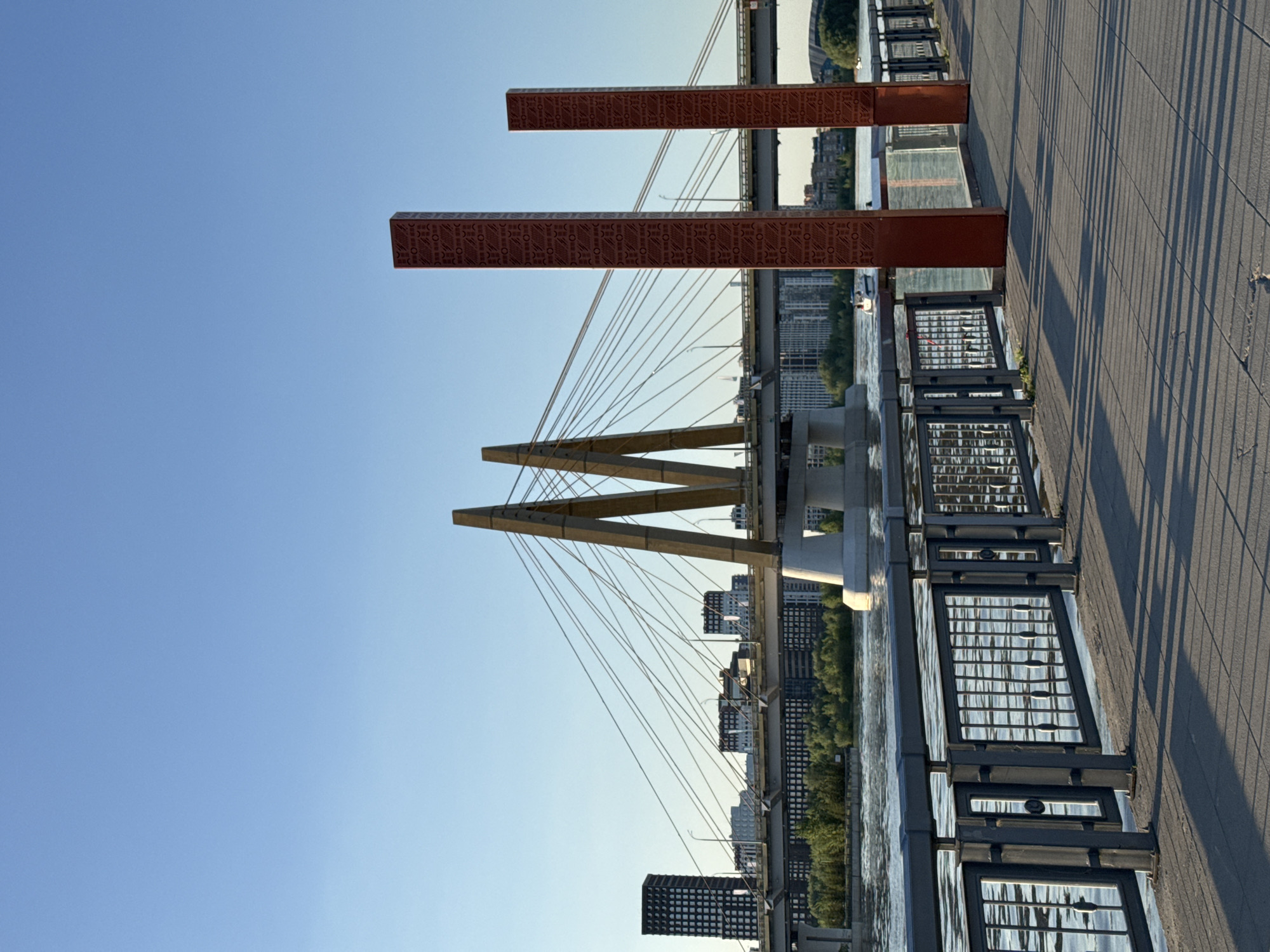 The image captures a view of the Millennium Bridge in Kazan, Russia. The bridge's distinctive "M" shaped pylon is visible against a clear blue sky. Suspension cables extend from the pylon towards the bridge deck. A pedestrian walkway borders the bridge, featuring decorative metal railings and reddish-brown rectangular columns with patterned designs. In the background, the skyline of Kazan includes modern buildings and green trees along the riverbank. The overall scene depicts a sunny, urban landscape centered around the Millennium Bridge.