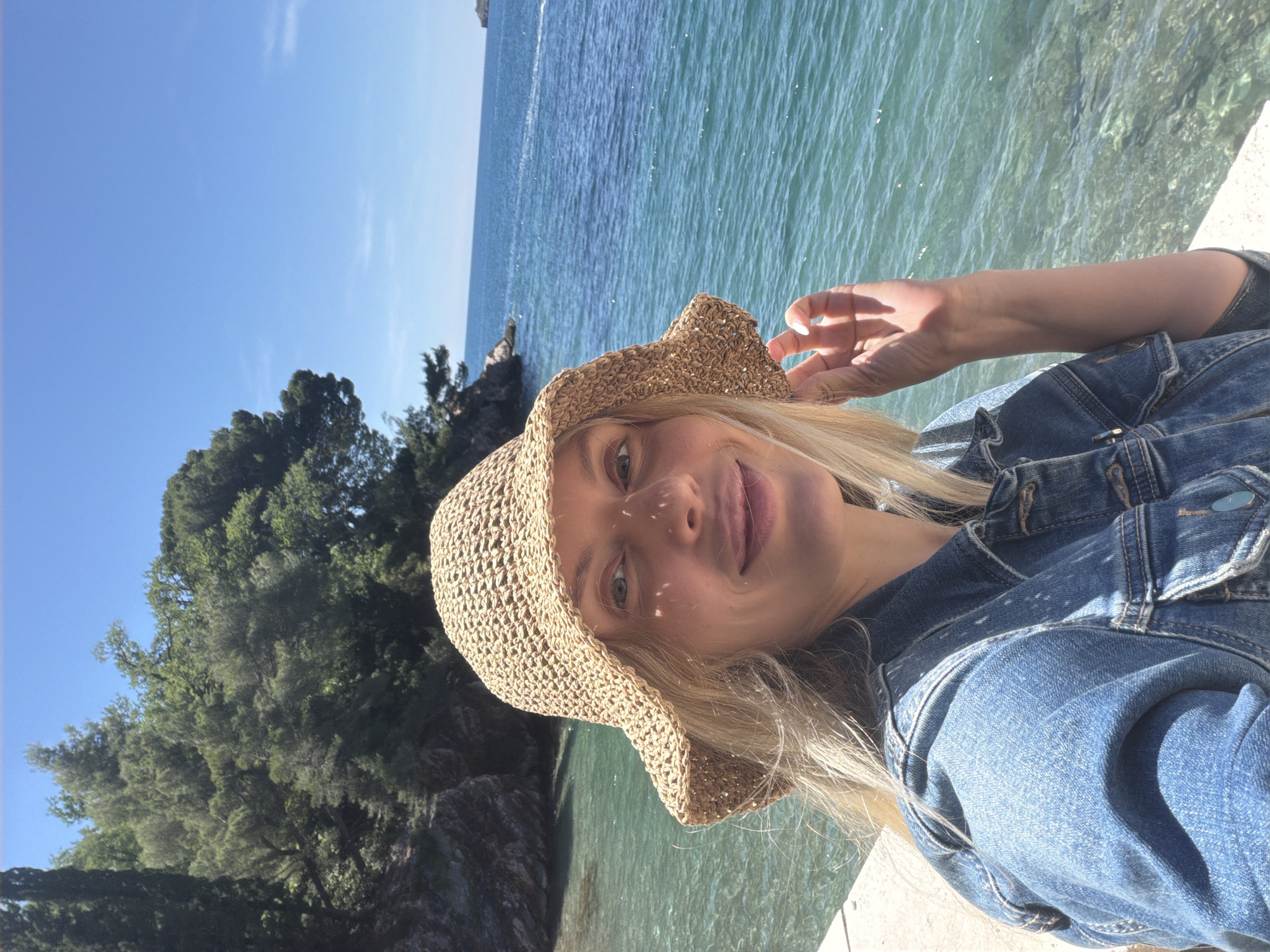 A medium shot depicts a blonde woman with fair skin wearing a woven sun hat and a denim jacket. She appears to be on a concrete ledge overlooking the turquoise waters of the Adriatic Sea, with an island densely covered in green trees in the background. The location is Sveti Stefan, Montenegro. The day is sunny and bright, casting light onto the woman's face.