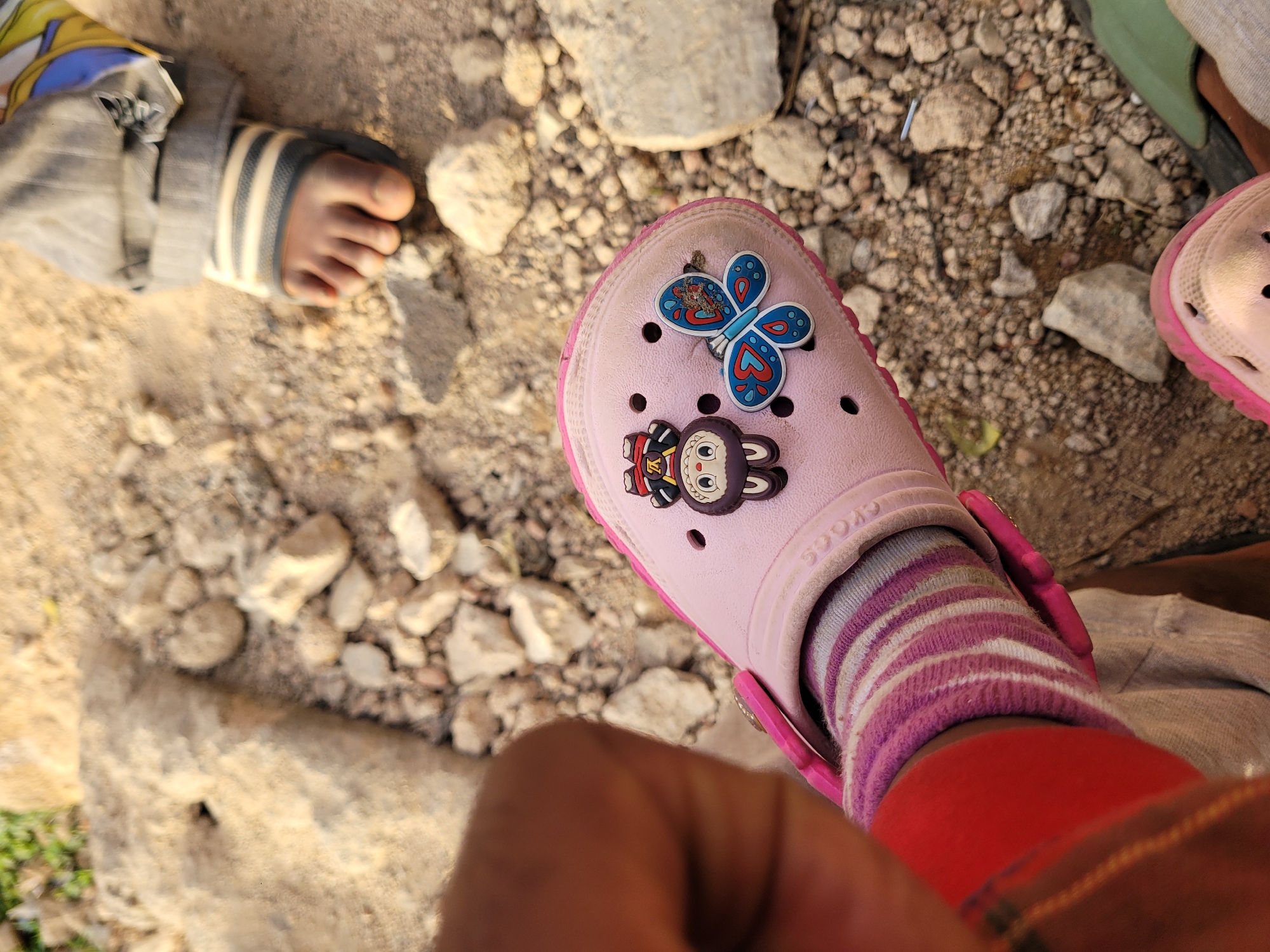 The image provides an overhead view of two children's feet on a dry, rocky outdoor ground in Barakin Akawo, Nigeria. In the foreground, a child's left foot is prominently featured, wearing a light pink Croc shoe. The shoe is personalized with two decorative charms: a blue butterfly with intricate patterns and a brown monkey-like character wearing a red shirt with a "VV" logo. The brand name "CROCS" is visible on the shoe's strap. Inside the Croc, the child wears a striped sock in shades of pink, purple, and white, with a solid red fabric piece (possibly part of clothing) visible around the ankle. To the left, another child's foot is partially visible, wearing a black sandal with white stripes and a grey and white striped sock, underneath what appears to be grey trousers or fabric. The ground is rough and uneven, covered with numerous small rocks, pebbles, and dirt, indicative of an unpaved area. The scene is brightly lit by natural daylight, suggesting it is daytime. No specific activity is depicted beyond the children standing or sitting on the ground.