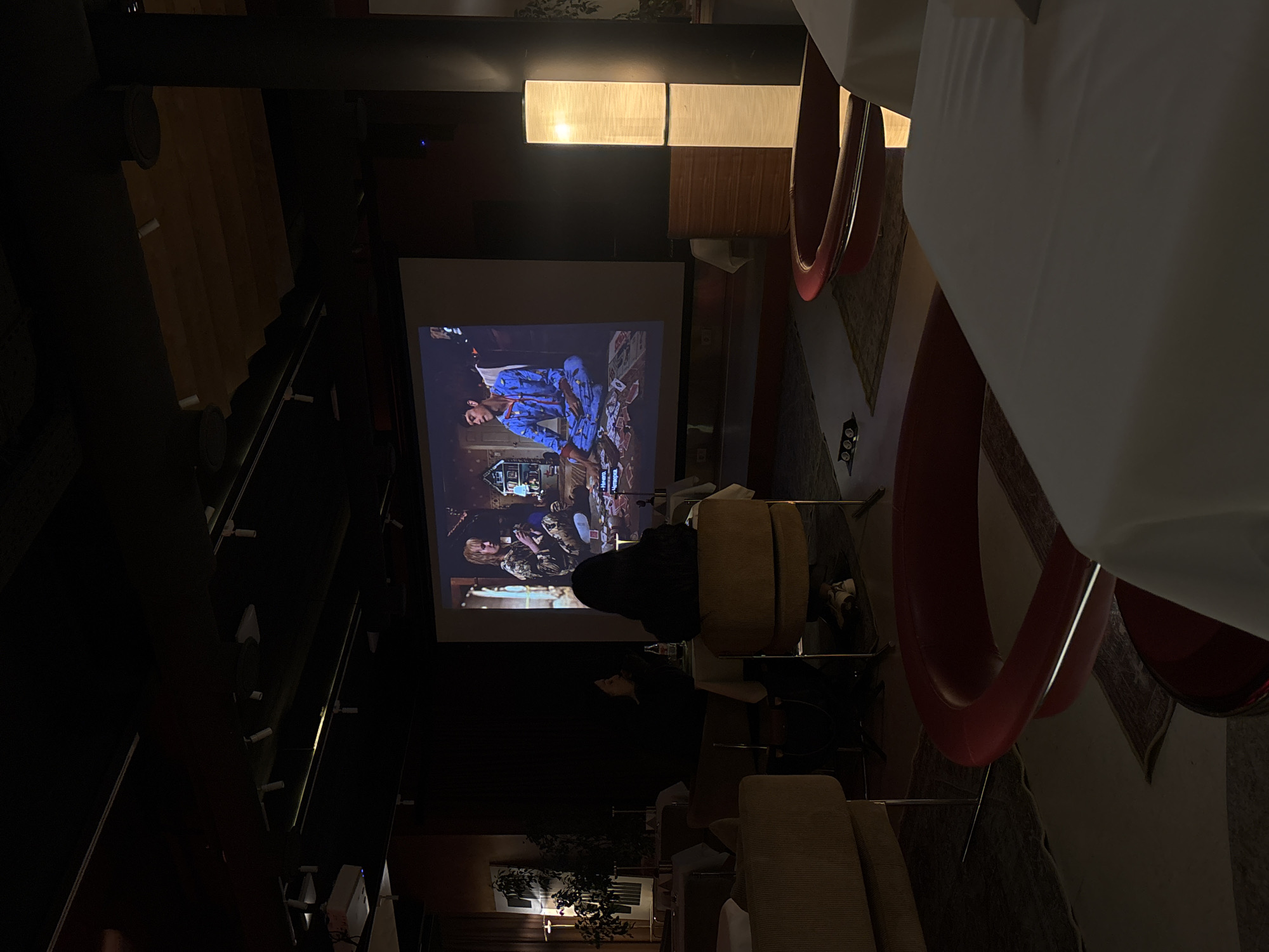 A gathering of people is watching a movie projected on a screen indoors. The scene is in Yerevan, Armenia. The projector is positioned to show the movie on a large screen. Several individuals are seated in chairs facing the screen, presumably watching the film. A light fixture illuminates the space near the screen. There are decorative plants. The seating includes both cushioned chairs and red, rounded chairs. A large white surface dominates the upper right portion of the image. A staircase is visible in the background.