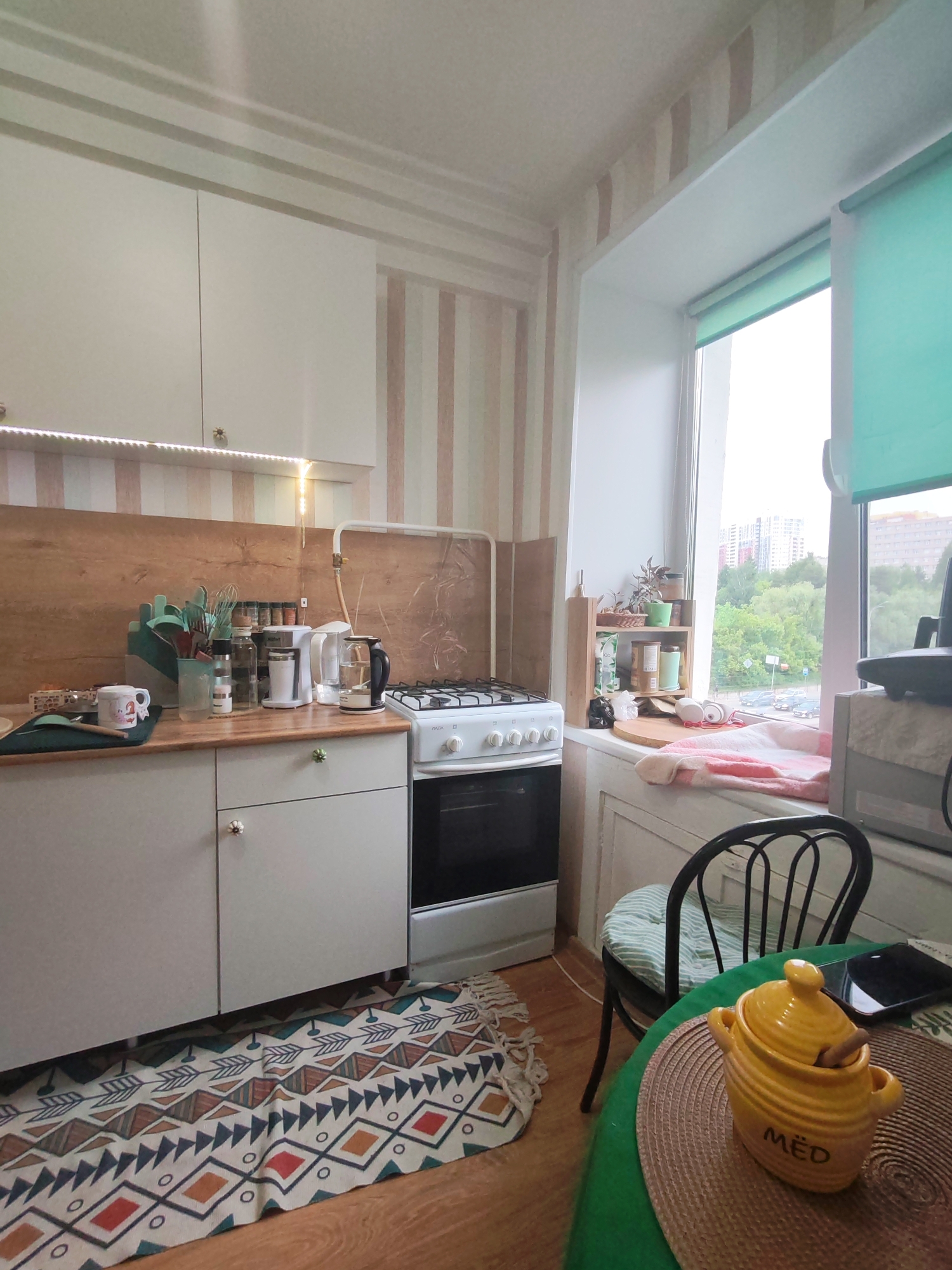 The image depicts a kitchen interior. A white gas range is situated next to light gray cabinets with decorative floral knobs. Above the cabinets are shelves and a countertop featuring various kitchen utensils, spices, and appliances like a coffee maker and kettle.
A window is visible on the right, showing an exterior view of Izhevsk, Russia with greenery and buildings. On the windowsill, a small wooden shelf unit holds plants and other items. A chair with a striped cushion is positioned near a table.
The table holds a yellow honey pot labeled "MËD" and a smartphone. An area rug with geometric patterns lies on the floor in front of the cabinets. The room is lit naturally through the window.