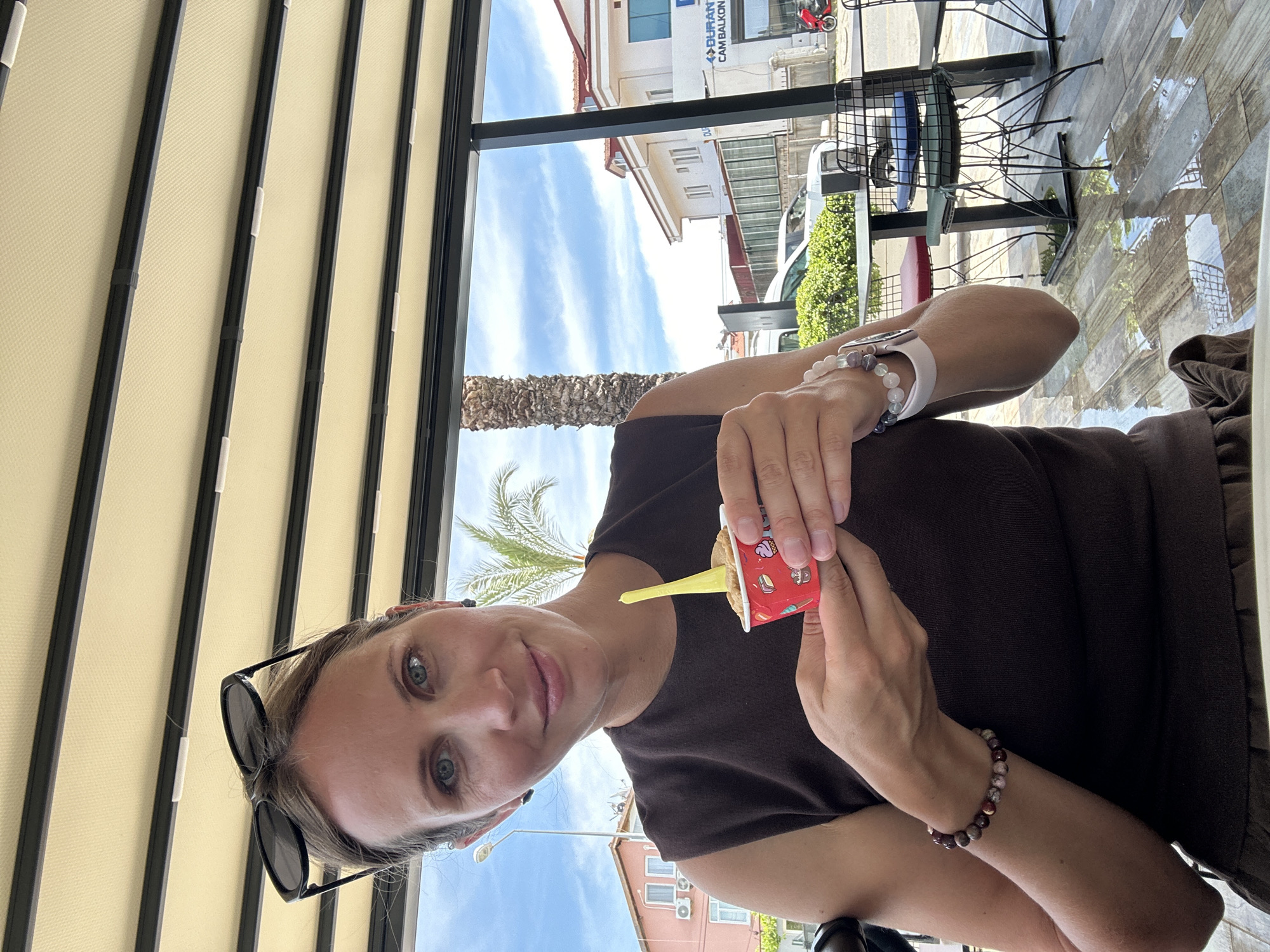 A person with fair skin, light brown hair, and blue eyes is holding a small container of ice cream with a yellow spoon. They are wearing a dark brown top and jewelry on their wrists. Sunglasses are perched on their head.
The setting appears to be an outdoor café in Fethiye, Turkey. Architectural elements of the city are visible in the background, including light-colored buildings, palm trees, and street furniture. The café has a retractable awning overhead. A car and a motorcycle are visible in the background. The table is made of tiles.