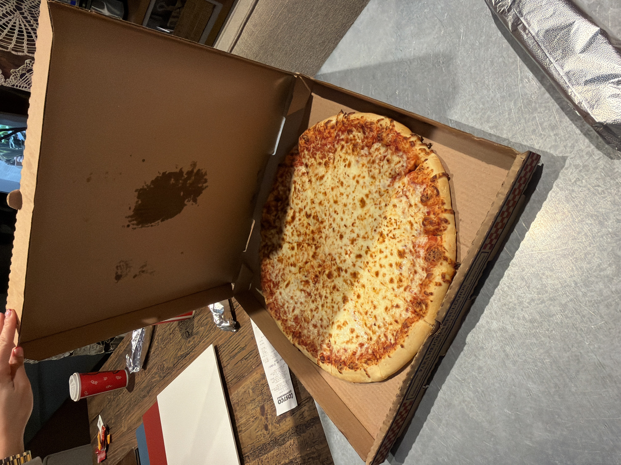 A partially opened pizza box contains a cheese pizza. The interior of the pizza box lid has a dark grease stain. The receipt shows a Costco logo, indicating this pizza was likely purchased from a Costco location. Based on the writing visible on the pizza box ("PARA MEJORES RESULTADOS"), the Costco location is likely in a Spanish-speaking region. Considering this information, the Costco is located in Puebla, Mexico. Part of a person's arm, wearing a gold watch, is visible in the lower left corner of the image. In the background, various objects and surfaces are visible, including a partially visible tabletop, a red paper cup, some foil wrappers, and a metal surface.