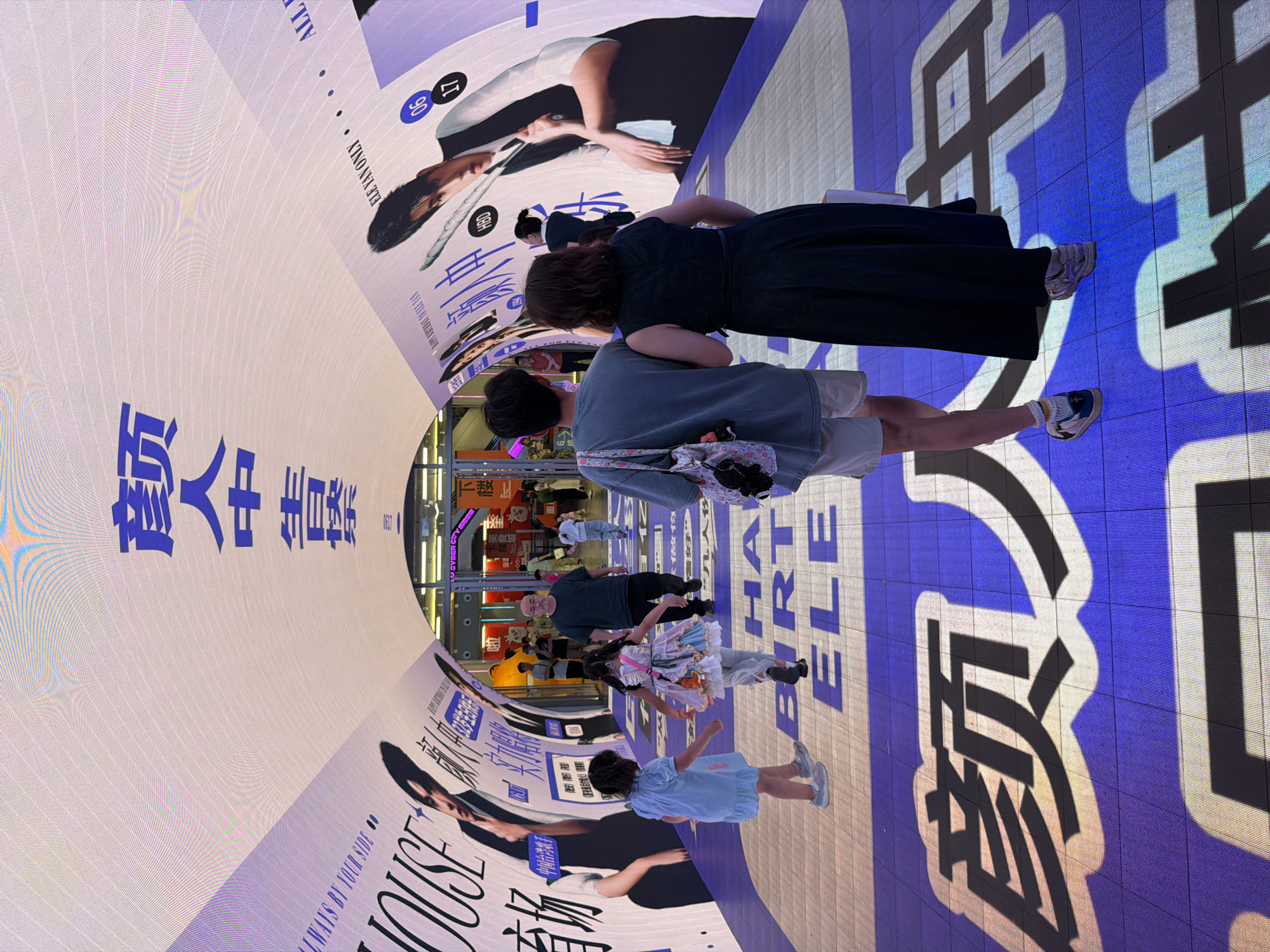The image shows a pedestrian tunnel in the Yuzhong District of China, illuminated by digital screens on the ceiling and walls. The digital display features large blue Chinese characters and images of a young man. On the floor of the tunnel, large blue Chinese characters are projected. Several people are walking through the tunnel. In the foreground, two young girls are walking away from the camera, followed by a man and a woman. Other people are visible in the background. The overall impression is a modern, urban public space with prominent digital advertising.