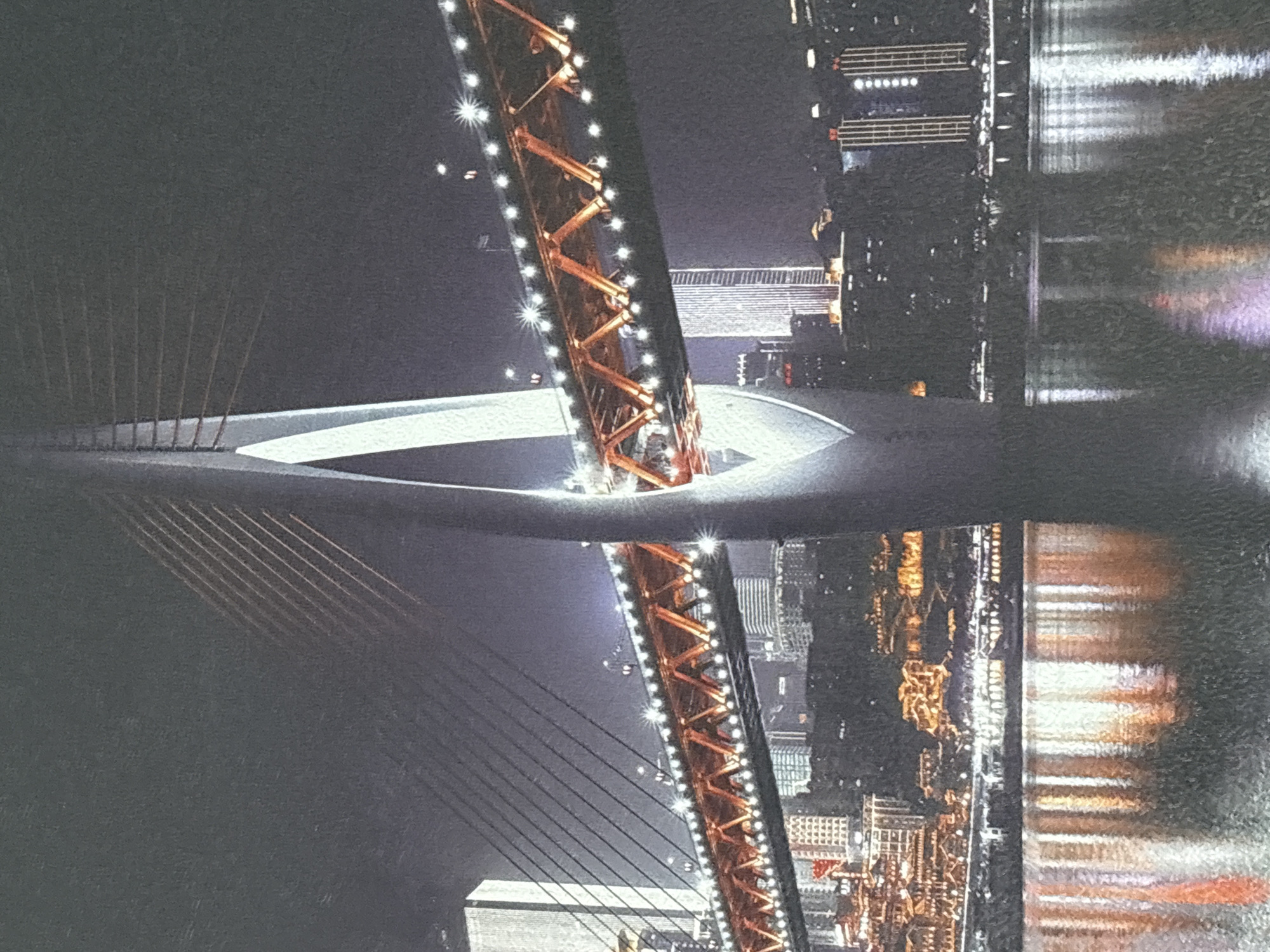 The media file presents a striking nighttime scene featuring a large, modern bridge spanning a body of water, with a vibrant city skyline on one side. The central element is a prominent bridge, likely a cable-stayed or suspension type, distinguished by its unique central pylon. This pylon is an intricately designed lattice or truss structure, glowing with an orange-golden hue and adorned with numerous bright white lights that create starburst effects. Dark support cables fan out diagonally from the pylon towards the bridge deck, which features a distinct white edge.
To the right of the bridge, a dense cityscape rises, illuminated by countless lights from buildings of various heights. The entire spectacle of the brightly lit bridge and the city lights is perfectly mirrored in the calm water below, creating a stunning and shimmering reflection that adds depth and visual appeal to the composition. The reflections in the water are clear, though slightly diffused, indicating still conditions.
The scene is clearly set at night, with a dark sky suggesting clear weather. No specific activity or event is depicted, but the image conveys a sense of modern urban grandeur and tranquility. No people are visible, and there is no discernible text within the image.