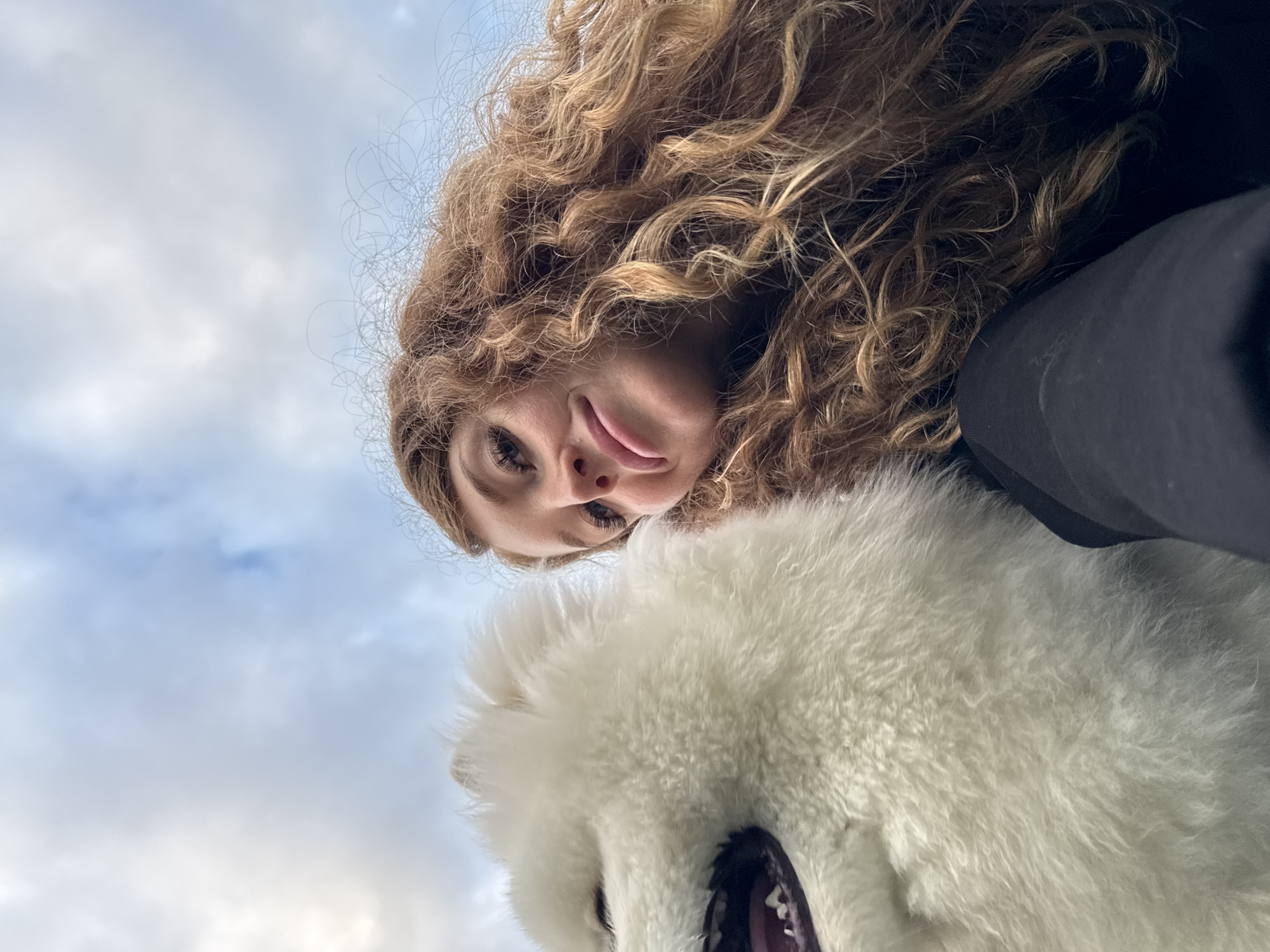 A low-angle, close-up, selfie-style shot shows a woman with long, curly, reddish-brown hair and a white fluffy dog. The woman is on the right side of the frame, leaning into the shot and smiling slightly. Her head is tilted, and her right eye is visible. The dog, a Samoyed, is on the left, with its mouth open, showing its teeth and tongue. The dog's head fills a significant portion of the left side of the image. The background is a cloudy, light blue sky, suggesting an outdoor setting. The image appears to have been taken in Chaikovsky, Russia.