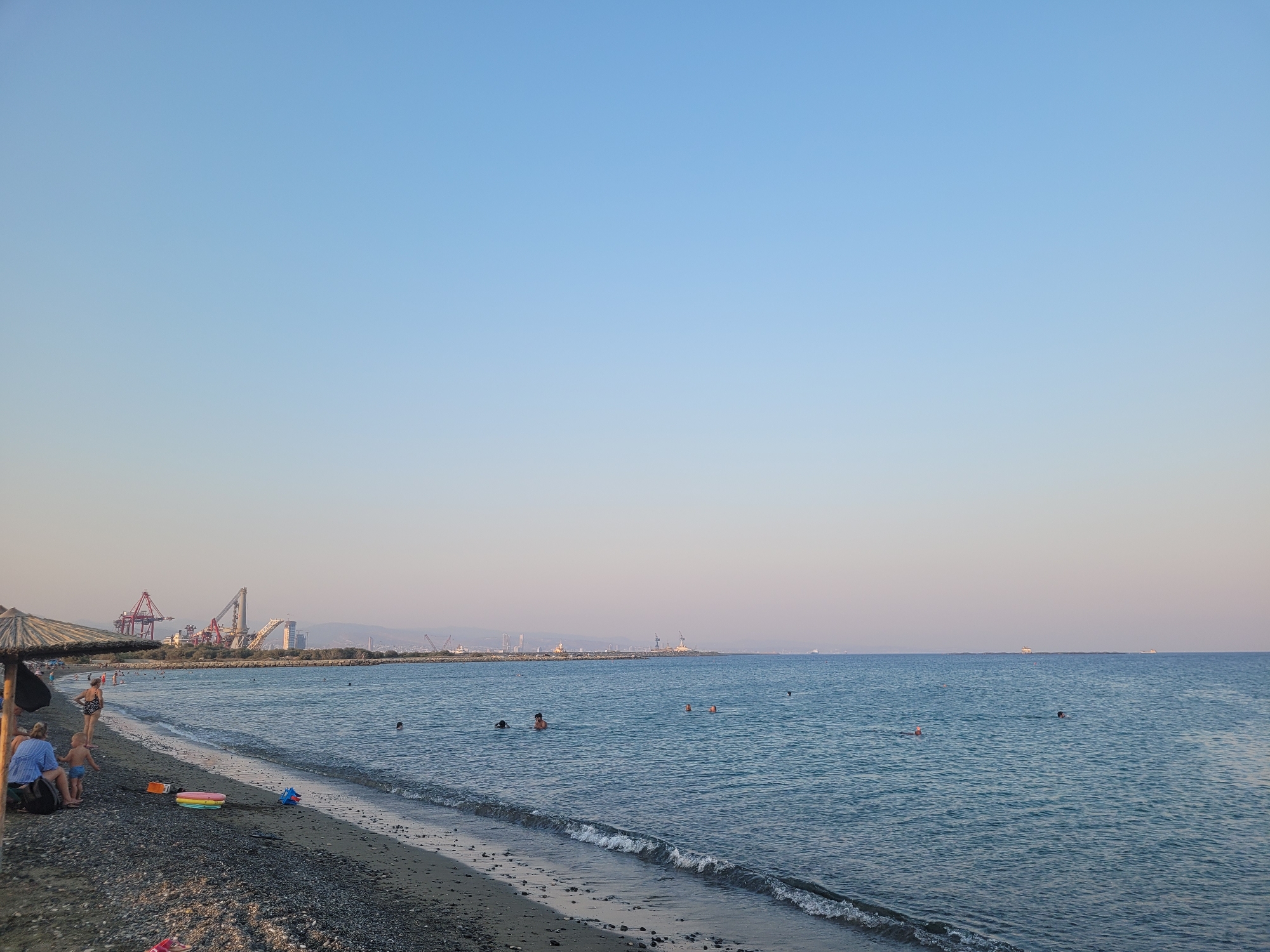 The image depicts a tranquil beach scene at Tserkezoi, Cyprus, under a clear sky. The foreground shows a pebbly beach with a few individuals relaxing. To the left, two adults are seated on the shore, one appearing to be looking at a mobile device. A small child stands near them. A thatched umbrella provides shade. In the water, several people are swimming and wading. The middle ground is occupied by the calm blue sea, with a gentle wave lapping at the shore. In the distance, a harbor area is visible with large cranes and industrial structures. The horizon is marked by a faint line of land and a vast expanse of the sea dotted with a few distant ships. The overall atmosphere is peaceful and serene, suggesting a late afternoon or early evening setting.