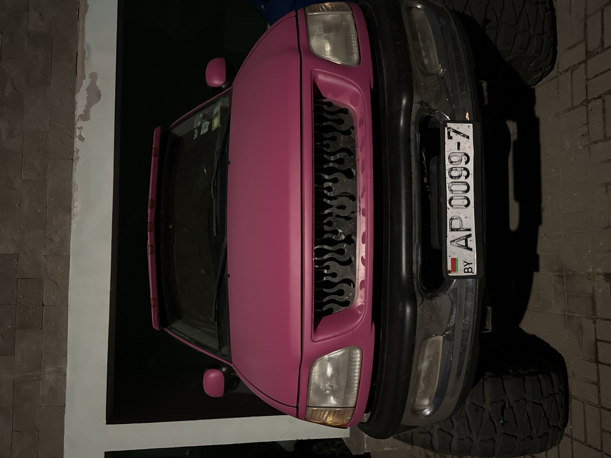 The image depicts the front of a pink vehicle parked on a brick-paved surface, potentially a driveway or parking area. The vehicle has a chrome bumper, large tires, and a distinctive grill with flame-like cutouts. The license plate is visible, displaying "AP 0099-7 BY," indicating it is a Belarusian license plate. The left side of the vehicle is situated next to a white wall and a darker brick wall, suggesting an enclosed space or building. The lighting conditions suggest it is nighttime. Minsk, Belarus is indicated by the vehicle license plate.