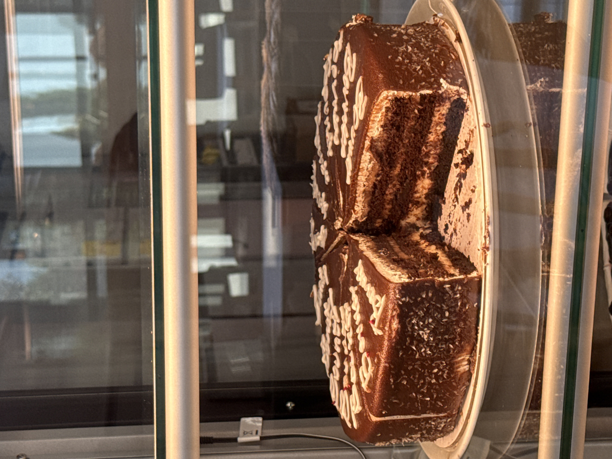 A cut chocolate cake with white icing and shredded coconut on the sides is displayed in a glass case. The cake has multiple layers of chocolate cake and cream filling. The icing on top is decorated with a swirly pattern. A slice has been removed from the cake. The background is blurred, but appears to be an indoor setting with reflections in the glass. The image is undated and the location is unknown.