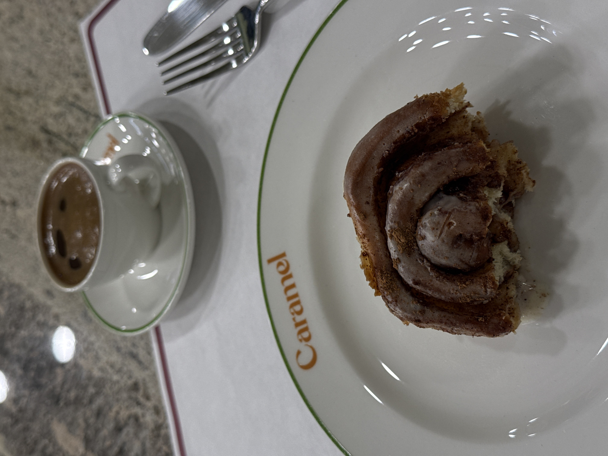 A cinnamon roll sits on a white plate with a green border, next to a cup of coffee and silverware. The word "Caramel" is printed on the plate. The location is Yerevan, Armenia. The table surface is speckled. The cinnamon roll appears to have been partially eaten.