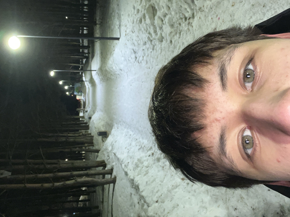 The image displays a close-up of a person's upper face in the foreground, positioned against a nighttime, snow-covered outdoor scene in Yelabuga, Russia. The person has brown hair with bangs, light brown/hazel eyes, and minor epidermal blemishes on the forehead. The background features a cleared, snow-covered pedestrian path extending into the distance, flanked by substantial snow banks. A dense line of tall, bare trees borders the left side of the pathway, with additional trees and snow visible on the right. The path is illuminated by a series of modern streetlights with bright, circular white light fixtures, casting light forward along the walkway. A dark waste receptacle is visible on the left side of the path, partially embedded in the snow. Further down the path on the right, a park bench is faintly discernible. In the far distance, indistinct lights and structures suggest an urban or recreational area. The scene depicts a winter environment at night.