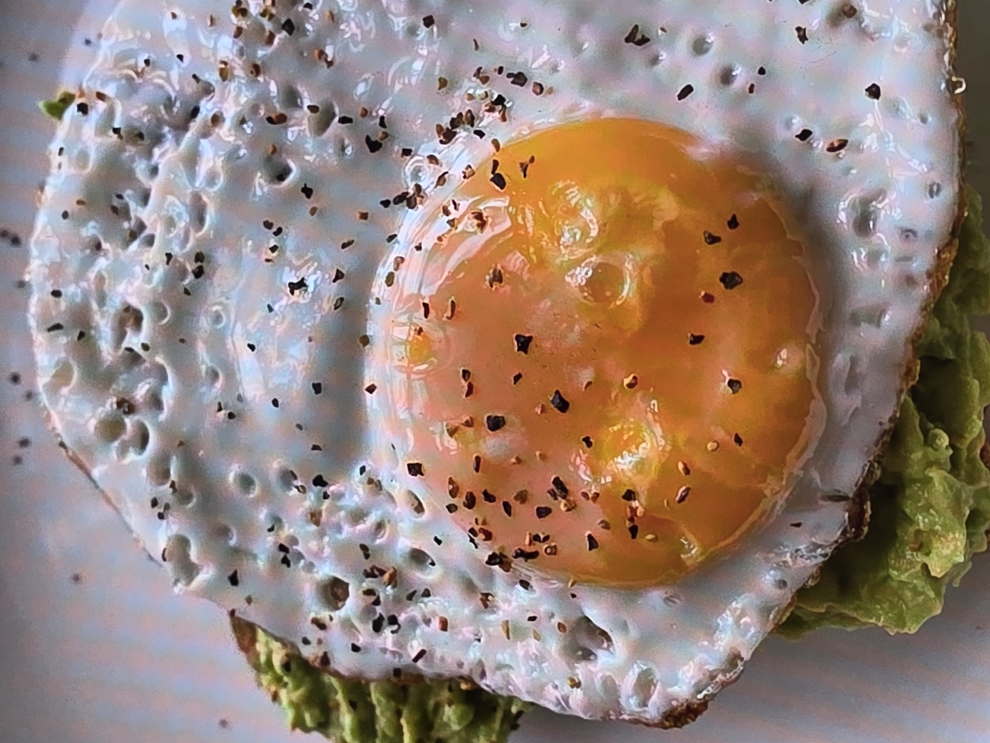 A close-up shot captures an overhead view of a fried egg with a runny yolk, seasoned with cracked black pepper, atop a bed of mashed avocado spread on a slice of toast. The avocado is a vibrant green, while the egg white is cooked to a crisp, showing small bubbles and brown edges. The yolk is a deep orange and appears to be slightly undercooked. The background is a neutral, light-colored surface, possibly a plate or tabletop. There is no indication of the city of Voluntari, Romania, or any environmental details from that location. The scene is focused solely on the prepared food item.